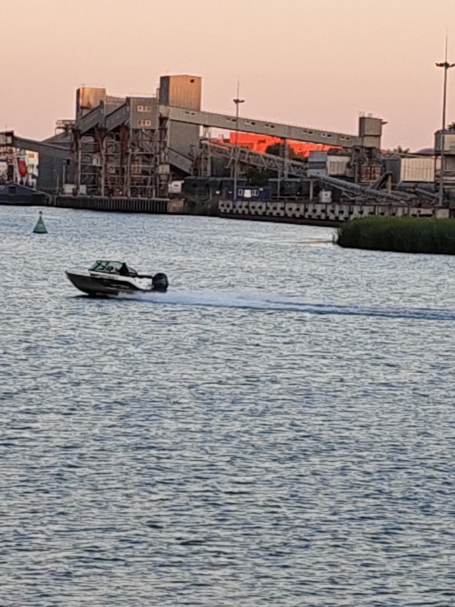 An eye-level, medium shot captures a motorboat speeding through the water, leaving a white wake behind it. The boat is silver and black with a black outboard motor. In the background, a large industrial facility with tall structures, some with orange accents, is visible along the waterfront. The sky above is a pale pink, suggesting either sunrise or sunset. The water is a deep blue-gray with ripples. A green buoy is visible in the foreground. The scene is an outdoor environment.