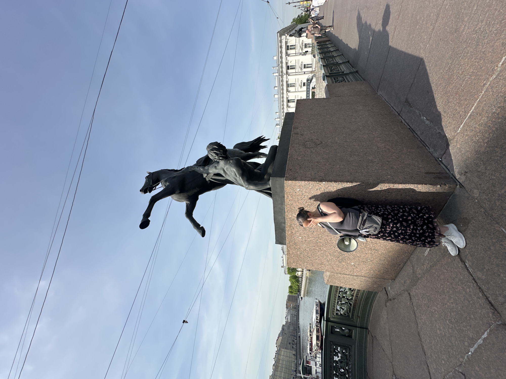 The image shows a bronze sculpture of a horse rearing up on its hind legs, held by a man with wings, possibly an angel, set atop a granite pedestal. The sculpture is located on a bridge in Saint Petersburg, Russia, with a view of the Neva River and cityscape in the background. Overhead wires crisscross the sky. A woman stands next to the pedestal, wearing a gray top, a floral-patterned skirt, and white sneakers, with a small bag slung across her chest and a megaphone resting on the ledge near her. The bridge appears to be made of large granite slabs. Other pedestrians can be seen further down the bridge.