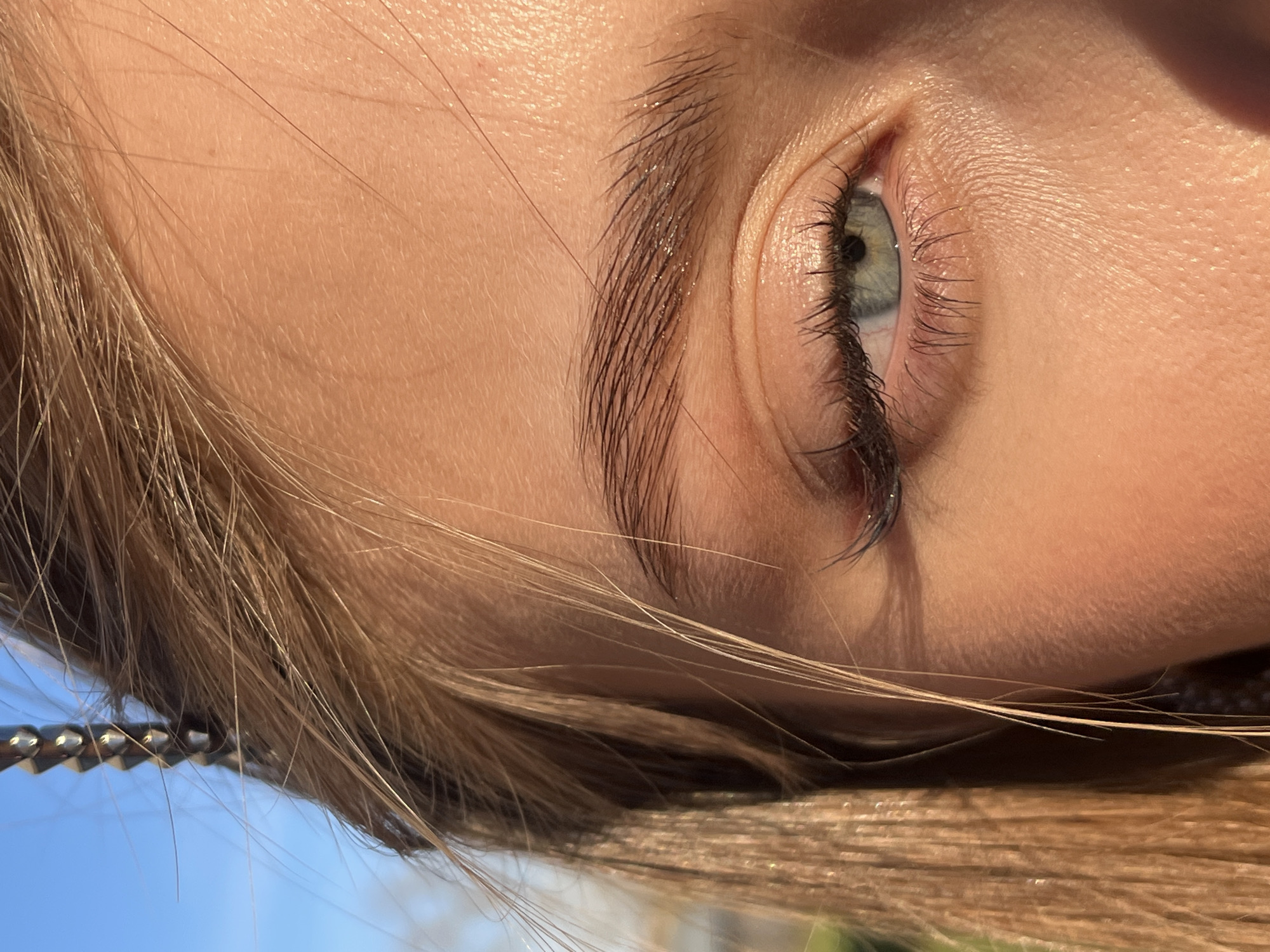 A close-up, tilted shot captures the eye and part of the face of a person with fair skin and light brown hair. The eye has a light blue iris and dark eyelashes. The eyebrow is dark brown and well-defined. A few strands of hair cross over the lower part of the face. In the blurred background, there are hints of blue sky and green foliage. A metal chain-like object is visible near the hair. The photo was not taken in Фоки, Russia, as there are no discernible location clues.
