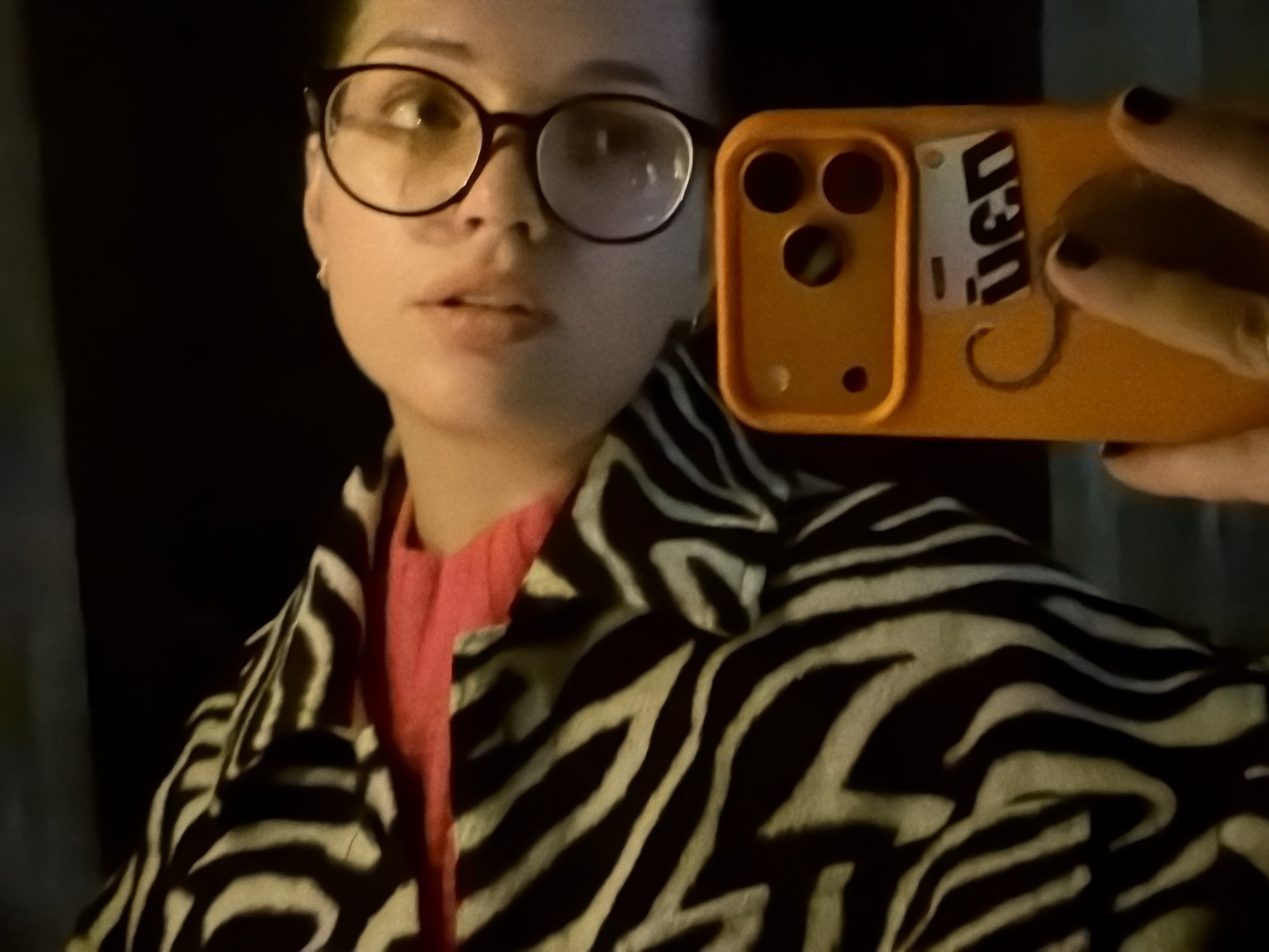 A person, likely female, is captured in a selfie taken indoors with low, artificial lighting. The subject wears dark-rimmed, round eyeglasses and has dark hair pulled back. They are dressed in a vibrant pink top beneath a prominent black and white zebra-print jacket or coat.
The person holds an orange smartphone in their right hand, positioned as if taking the photo. The phone case features cutouts for what appear to be three camera lenses, suggesting an iPhone model. A white rectangular sticker with dark text reading "üCD" is affixed to the back of the phone case. Dark nail polish is visible on the fingers holding the phone.
The background is uniformly dark and indistinct, providing no specific setting details or architectural cues. The person's gaze is directed slightly upwards and to the side, consistent with looking at a phone screen while taking a selfie. The lighting creates reflections on the glasses and illuminates the subject's face. The overall mood is casual, with a neutral expression. The provided location context of Большая Шильна, Russia, is not visually discernible within the image.