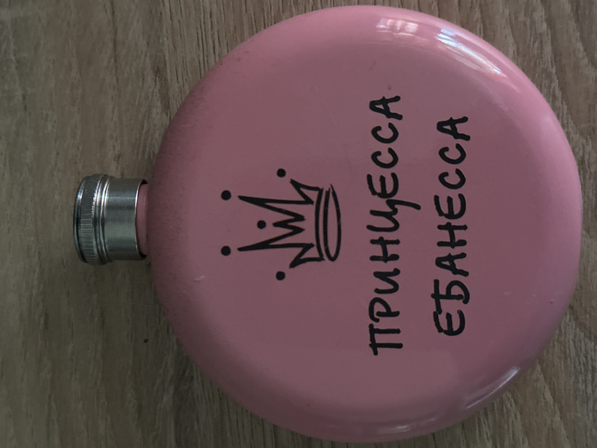 A pink flask with a screw-on cap is lying on a wooden surface. The flask is decorated with a black crown and the Russian words "ПРИНЦЕССА ЕБАНЕССА" written vertically on the right. The words translate to "Princess Crazy." The location in Zelenograd, Russia, is not discernible from the image.
