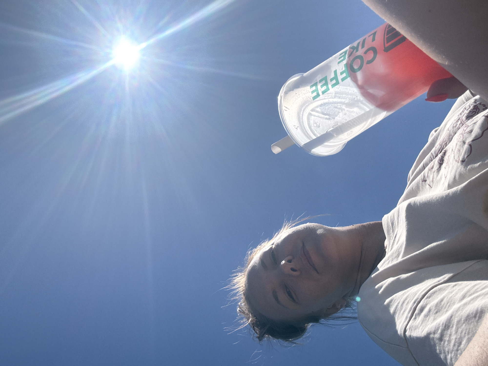 A low-angle, outdoor, eye-level shot shows a woman in the foreground holding a clear plastic cup with a red beverage and a straw. The cup has green text that reads "COFFEE LIKE". The woman is wearing a light-colored t-shirt. Her hair is pulled up, and some strands are sticking out. Her face is looking towards the camera. In the background, the sky is bright blue with the sun creating a lens flare effect. The image does not provide any specific details to determine the location in Dmitrov, Russia.