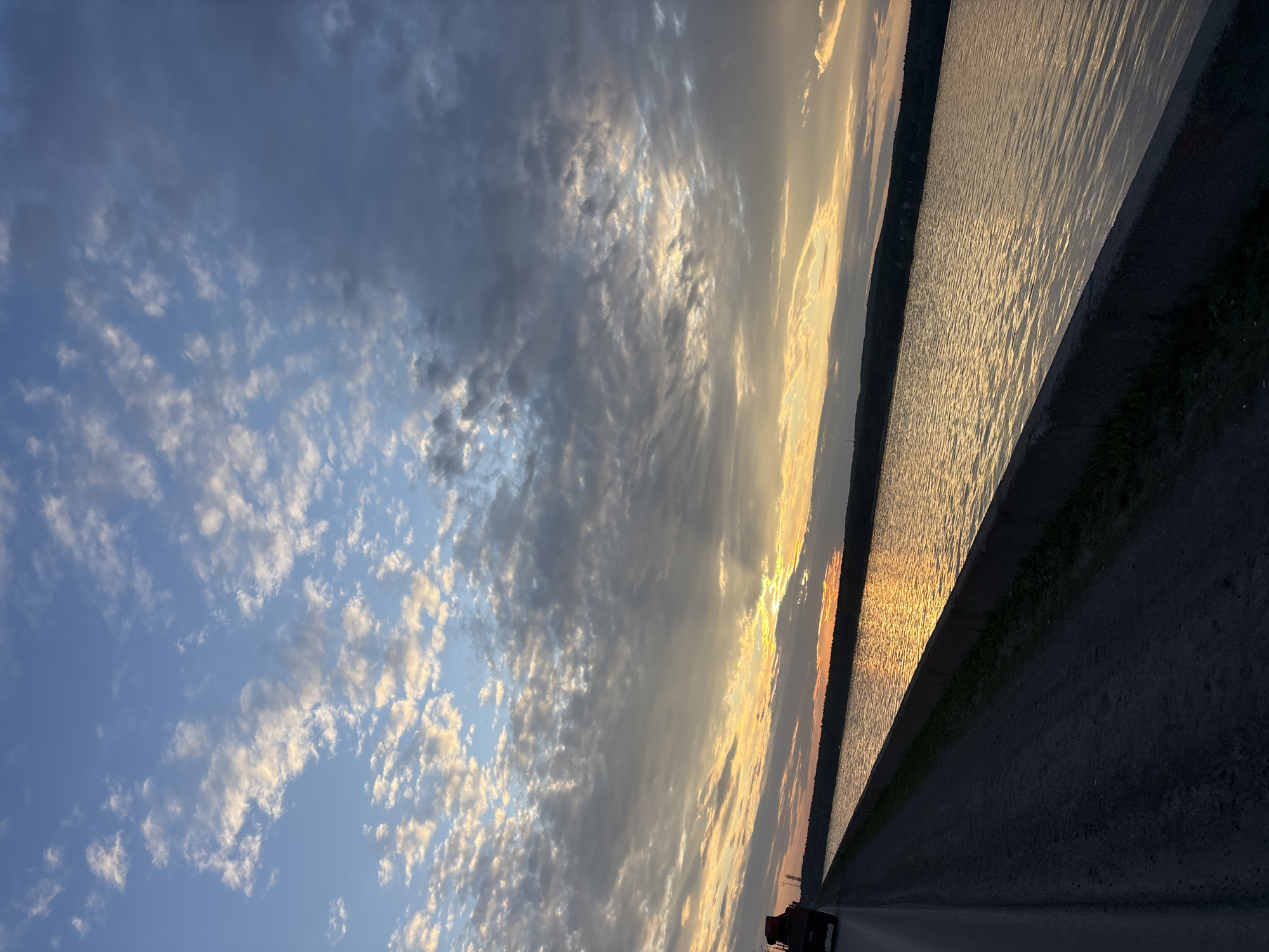 A body of water, possibly the Kama River, is seen reflecting the golden hues of a sunset. The water is bordered by a concrete embankment and a paved road. A dark-colored vehicle, potentially a truck, is partially visible on the road, moving away from the viewer. The sky is filled with a mix of clouds, with some areas illuminated by the setting sun and others appearing darker and more ominous. The overall scene suggests a tranquil, natural setting at dusk, with a hint of human presence from the road and vehicle. The location is identified as Chaikovsky, Russia.