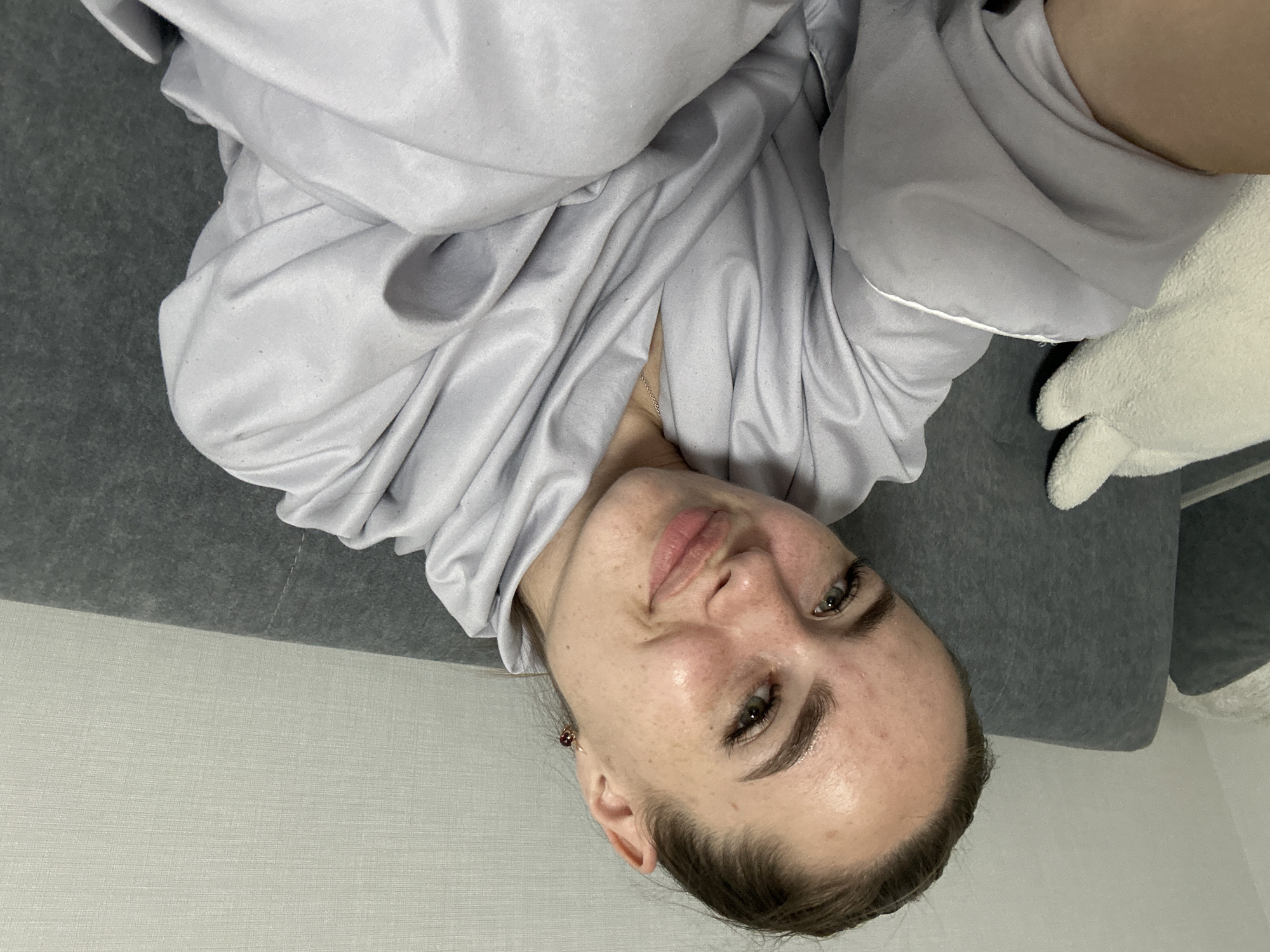 An unidentifiable woman with light skin and freckles, wearing a light gray robe, rests on a gray sofa. Her hair is pulled back into a bun, and she has noticeable makeup, including eyeliner and mascara. A white plush object, possibly a stuffed animal, is visible to her left. The background consists of a textured off-white wall. The lighting suggests an indoor setting, and the overall mood is relaxed. The image is a close-up, focusing on the woman's face and upper body.