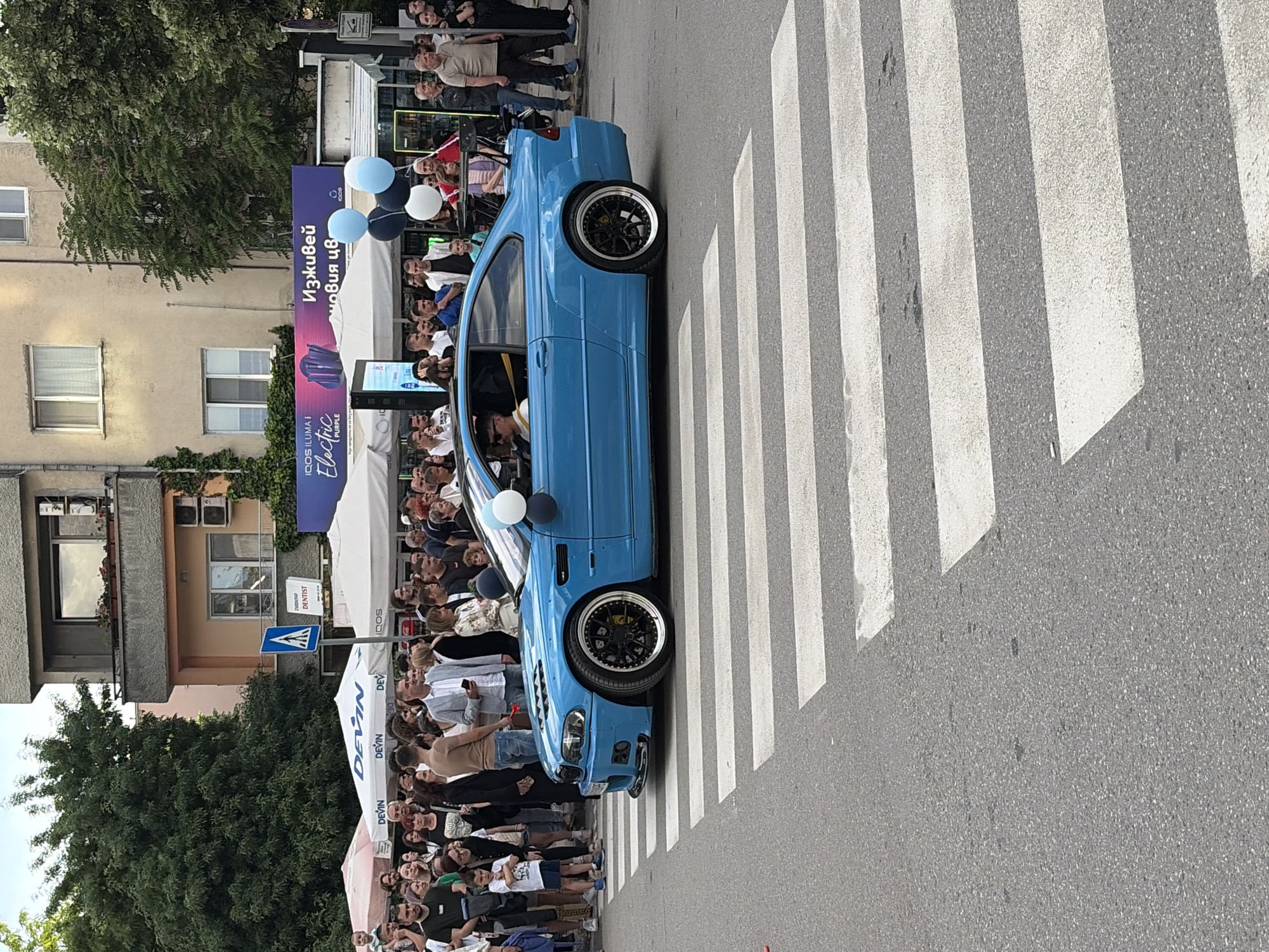 The scene is an outdoor event in an urban setting, seemingly a city street in Unknown, Unknown. A bright blue modified car is parked diagonally across a zebra crossing, effectively blocking pedestrian traffic. The car has black rims and what appear to be aftermarket modifications.
A large crowd of people of varying ages and ethnicities is gathered on both sides of the car, most likely observing or participating in the event. A few people are standing on the crosswalk next to the vehicle.
In the background, there are buildings, including a multi-story building with windows, balconies, and air conditioning units. A blue and purple sign can be seen, partly obscured, displaying the text "IQOS ILUMA I Electric Purple." Further back, there are white tents and signage with the word "Devin" visible. Balloons in white and light blue are present near the car. A blue pedestrian crossing sign is visible near the tents. The environment suggests a public event or display, possibly related to automobiles or the promoted brands.
