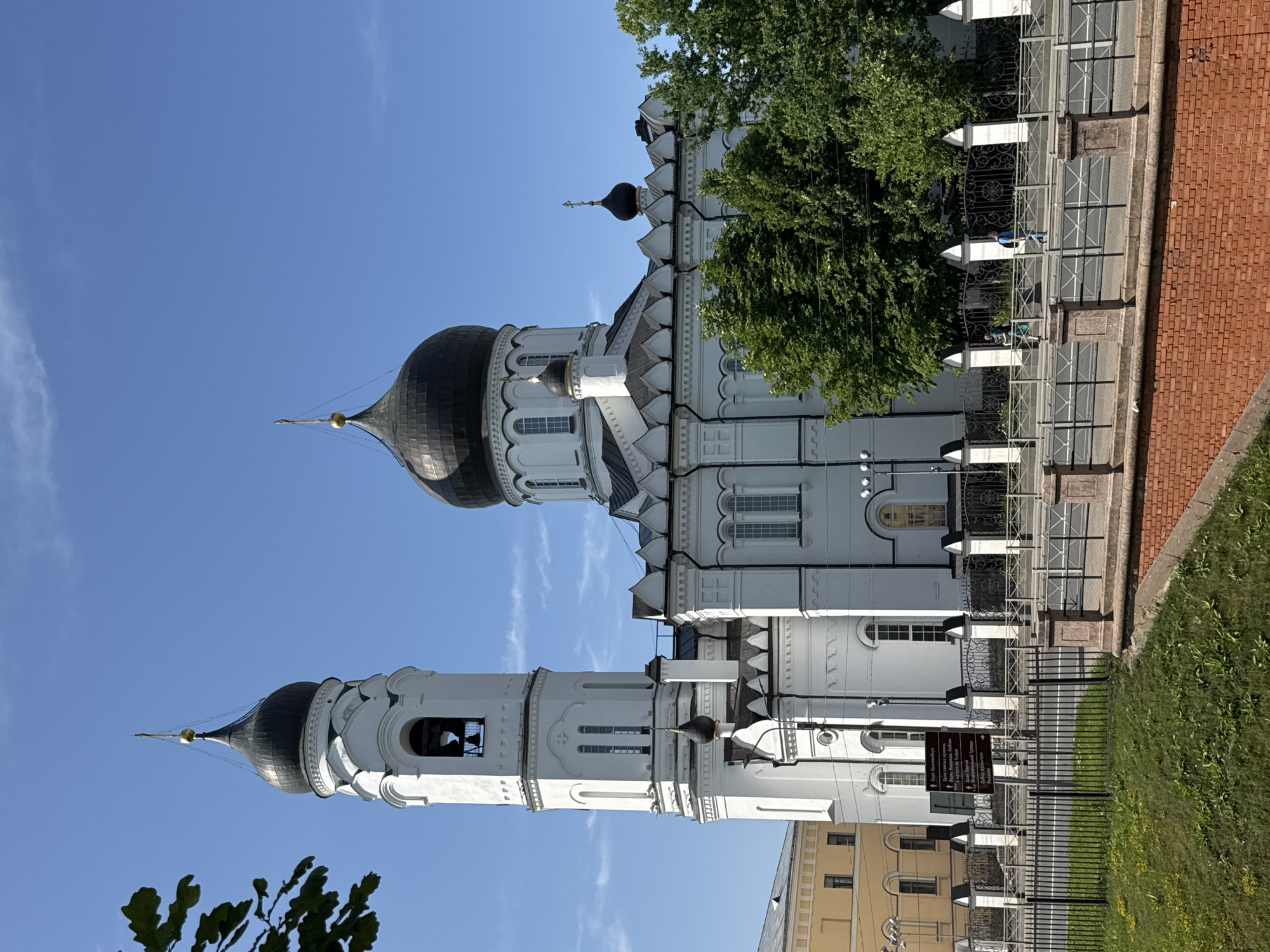 The image captures a bright, sunny day in Lomonosov, Russia, with a clear blue sky overhead. The primary subject is a white Orthodox church with a tall, multi-tiered bell tower and a prominent black dome. The architecture features intricate white decorative elements and arched windows. A smaller, dark dome topped with a cross is visible on a section of the church.
To the right of the church, a lush green tree provides a vibrant contrast to the white facade and blue sky. A metal fence with ornate detailing separates the church grounds from a pathway and a red brick wall. A grassy area with scattered yellow flowers borders the fence, indicating a well-maintained outdoor space.
In the background, a section of a yellow building is visible, suggesting an urban or historical setting. The orientation of the image is portrait, and it appears to have been taken from a slightly elevated viewpoint, looking upwards towards the church. There are a few hints of people near the fence, but they are not clearly discernible. A sign with Cyrillic text is partially visible near the fence, but the text is too small to read clearly. The overall impression is one of a serene and beautiful religious site on a pleasant day.
