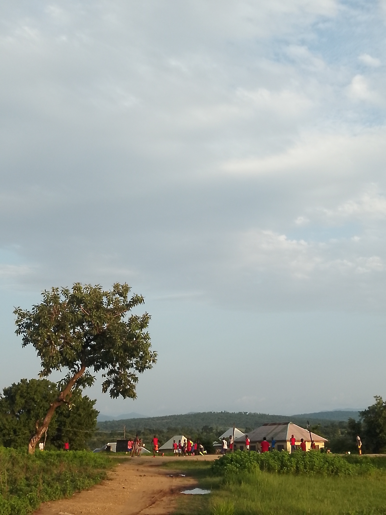 In Serti, Nigeria, an outdoor scene unfolds under a vast, cloudy sky. A dirt road curves through a grassy landscape, bordered by lush vegetation and a large, leafy tree on the left. In the middle ground, several small buildings, possibly tents or simple dwellings, are set up. A group of people, predominantly wearing red shirts, are gathered around these structures, with some appearing to be interacting or engaging in activities. Further in the distance, rolling hills create a natural backdrop to the settlement. The overall impression is one of a rural community, perhaps during an event or gathering, captured in natural light with a slightly overcast sky.