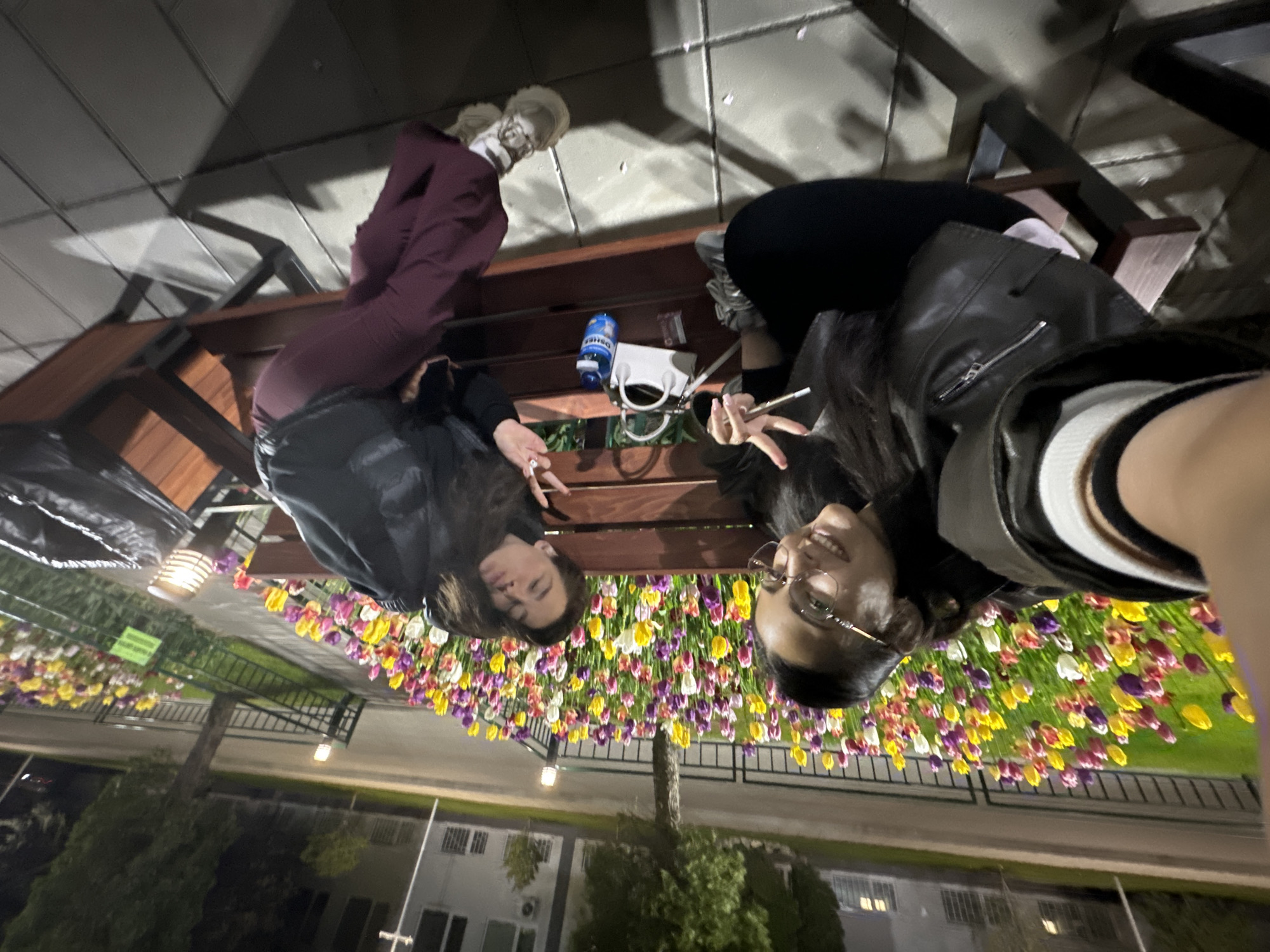 Two women sit on a wooden bench outdoors at night in Sofia, Bulgaria, with a dense flower display of multi-colored tulips behind the bench. The woman on the left wears maroon pants, a dark jacket, and light-colored shoes. The woman on the right is dressed in black pants and a black leather jacket. She wears glasses and smiles towards the camera, holding a cigarette. On the bench between them, there is a white purse and a blue water bottle. In the background, there are buildings, trees, streetlights, and a metal fence that surrounds the flowers.