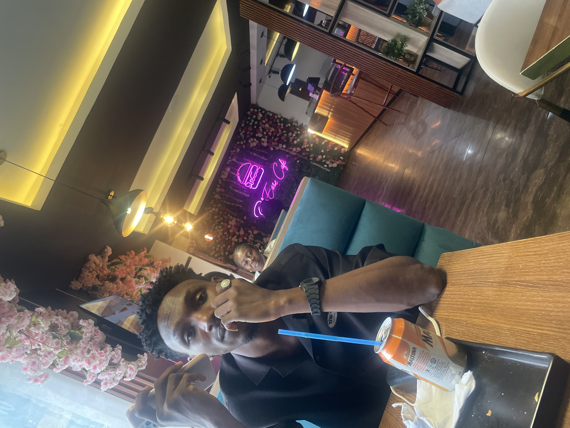 A male, presumably in his twenties, is seated at a wooden table in a cafe-style establishment. He is wearing a black shirt and has a dark complexion and short, curly hair. He is looking at the camera with a beverage can of "Maltina" with a blue straw in front of him, on a small black tray. He is holding a mobile phone in his left hand and the right hand is raised to his face, with a ring visible on his finger. Another person, presumably a female, is visible in the background, seated on a turquoise bench. The background includes a floral arrangement, a neon sign, and interior design features of the cafe. The image seems to have been taken at a restaurant in Adankolo, Nigeria.