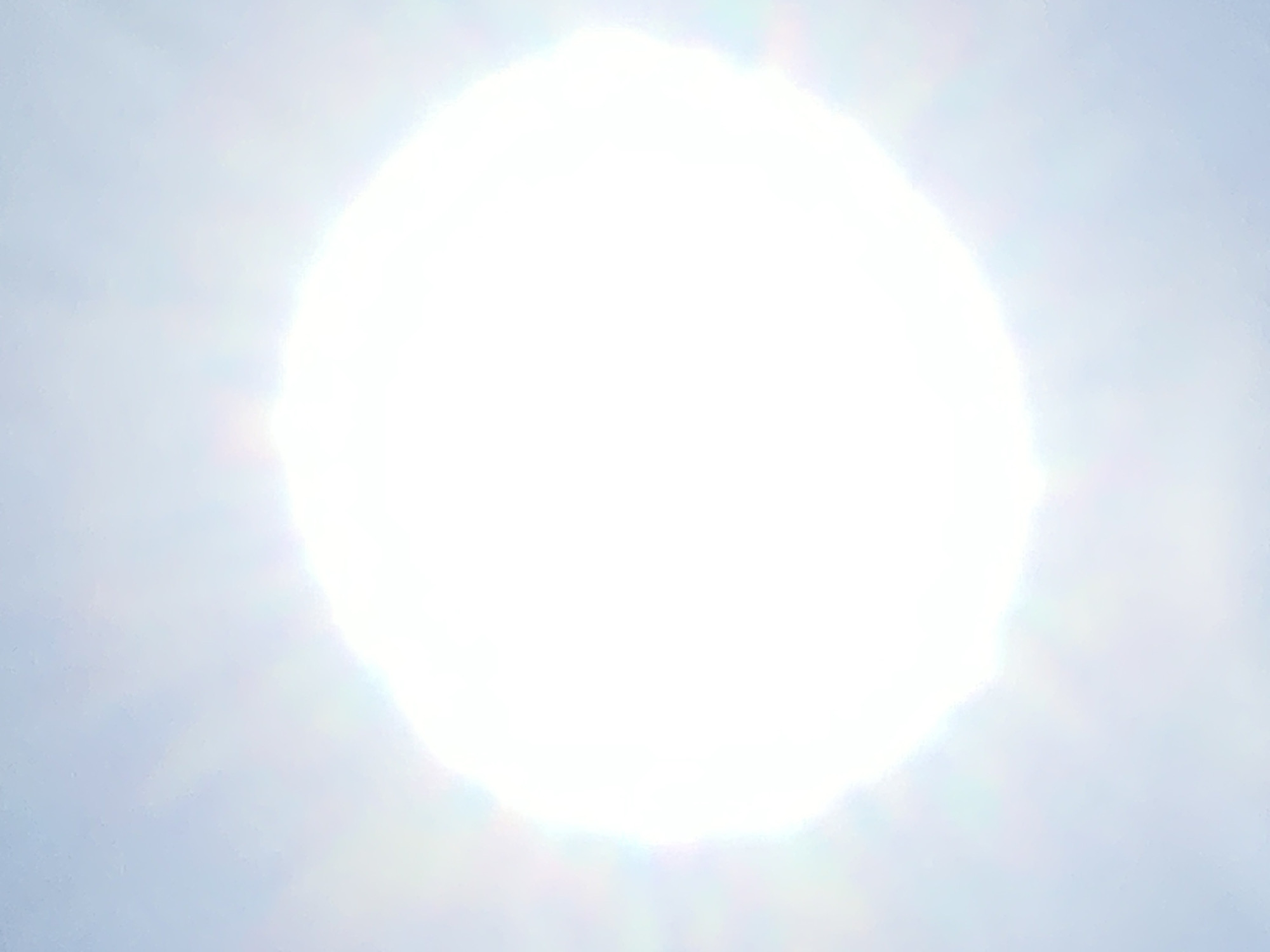 The image shows a bright, spherical object in the sky, likely the sun, surrounded by a hazy or cloudy atmosphere. The light is intense, creating a lens flare effect with a soft, diffused glow. There are subtle rainbow-like refractions of light visible around the edges of the bright object. The background is a pale blue, suggesting an outdoor daytime scene. There are no visible landmarks or elements that would indicate a specific location within the city of Kolomyia, Ukraine. The image is a close-up of the sky and the bright celestial body.