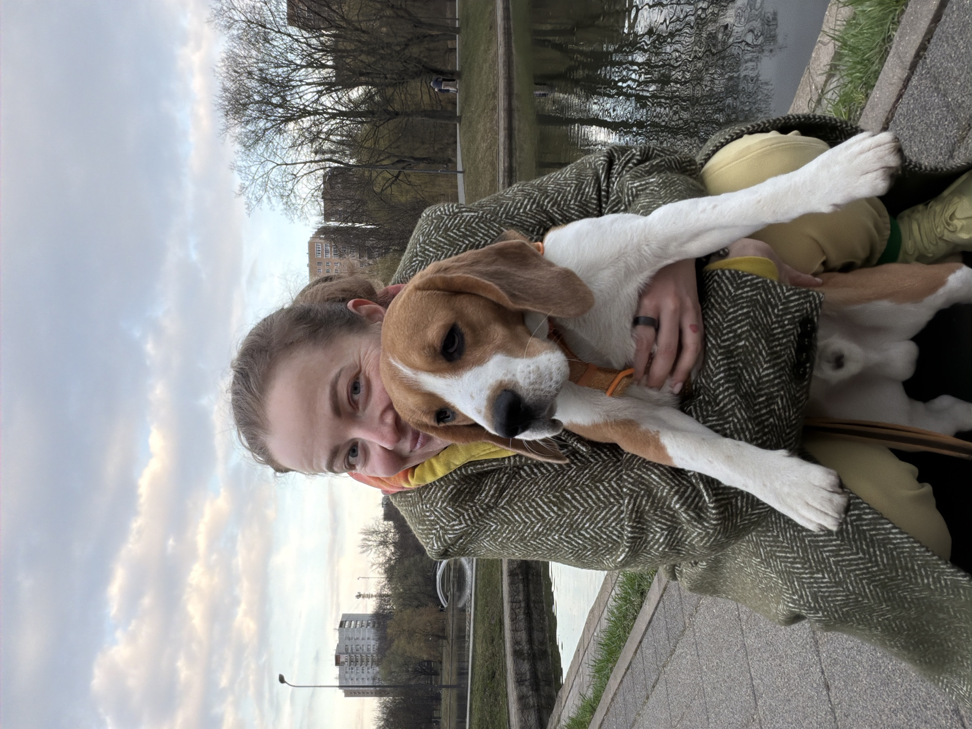 A woman is holding a dog, possibly a beagle, in her arms. She is wearing a green herringbone coat over a yellow sweatshirt. They are outdoors next to a body of water reflecting trees and the sky. Buildings are visible in the background. Based on the architectural style and the presence of a distinctive fountain, the location appears to be in Moscow, Russia, likely near a park with a pond or canal.