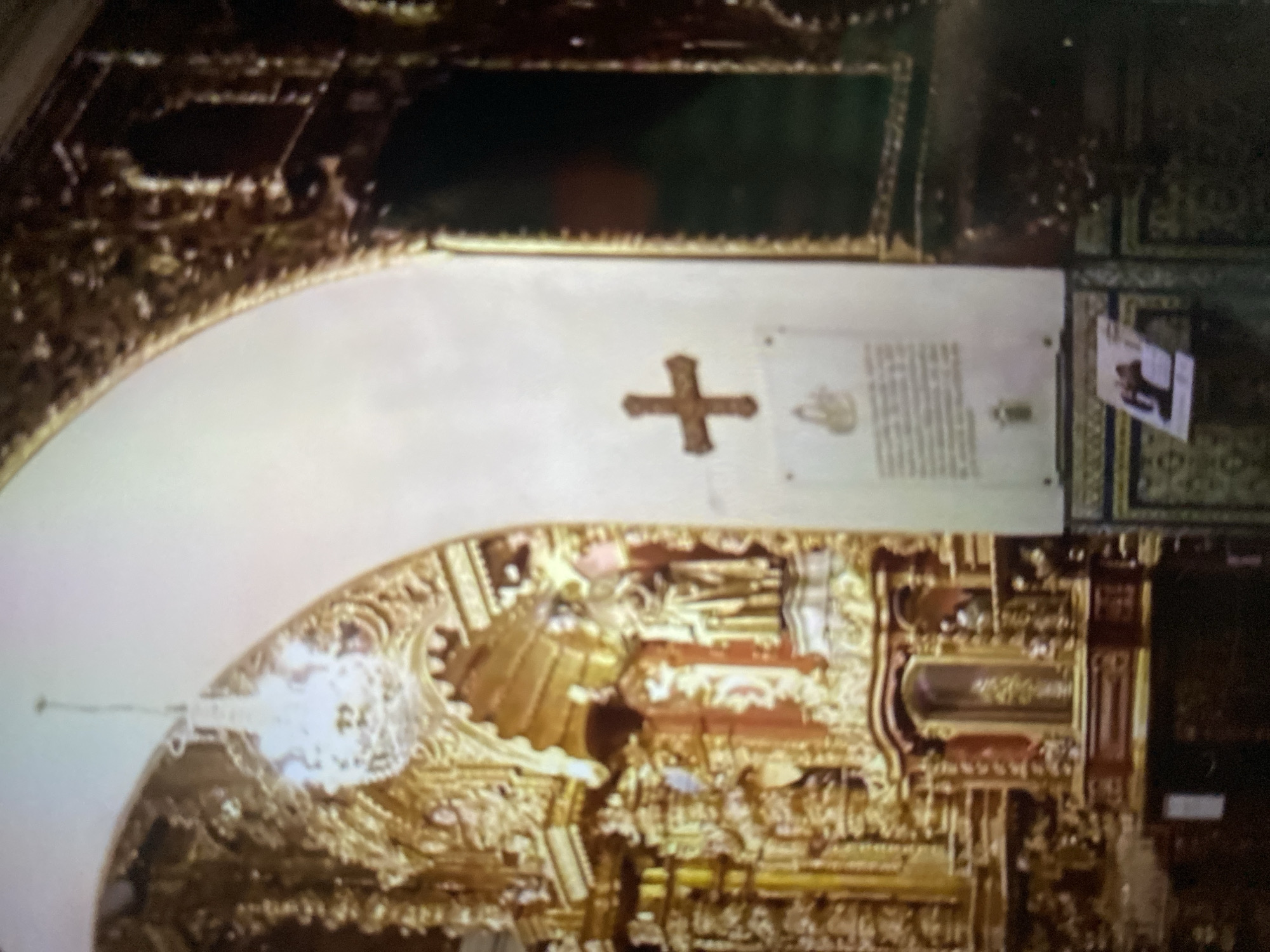 The image shows the interior of a church in Tinaquillo, Venezuela. The church features a white arched ceiling with ornate golden details. On the wall is a simple brown cross, a plaque, and a small photo. Below is a complex golden structure with niches, likely an altar or reredos. A chandelier hangs from the ceiling.
