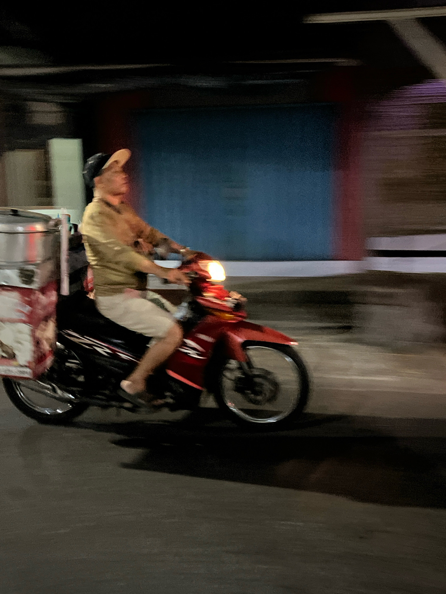 A person wearing a tan long-sleeved shirt, light-colored shorts, and a tan baseball cap rides a red scooter with a large silver container attached to the back. The container has a red and white patterned wrapping around its base. The person is in motion, indicated by the motion blur of the background. The scooter has a visible headlight and tire. The scene takes place on a dark, paved road, possibly at night, with indistinct building structures in the background, one of which appears to be a blue wall. The overall impression is a street scene in Banguntapan, Indonesia, with a delivery or vendor on a scooter.