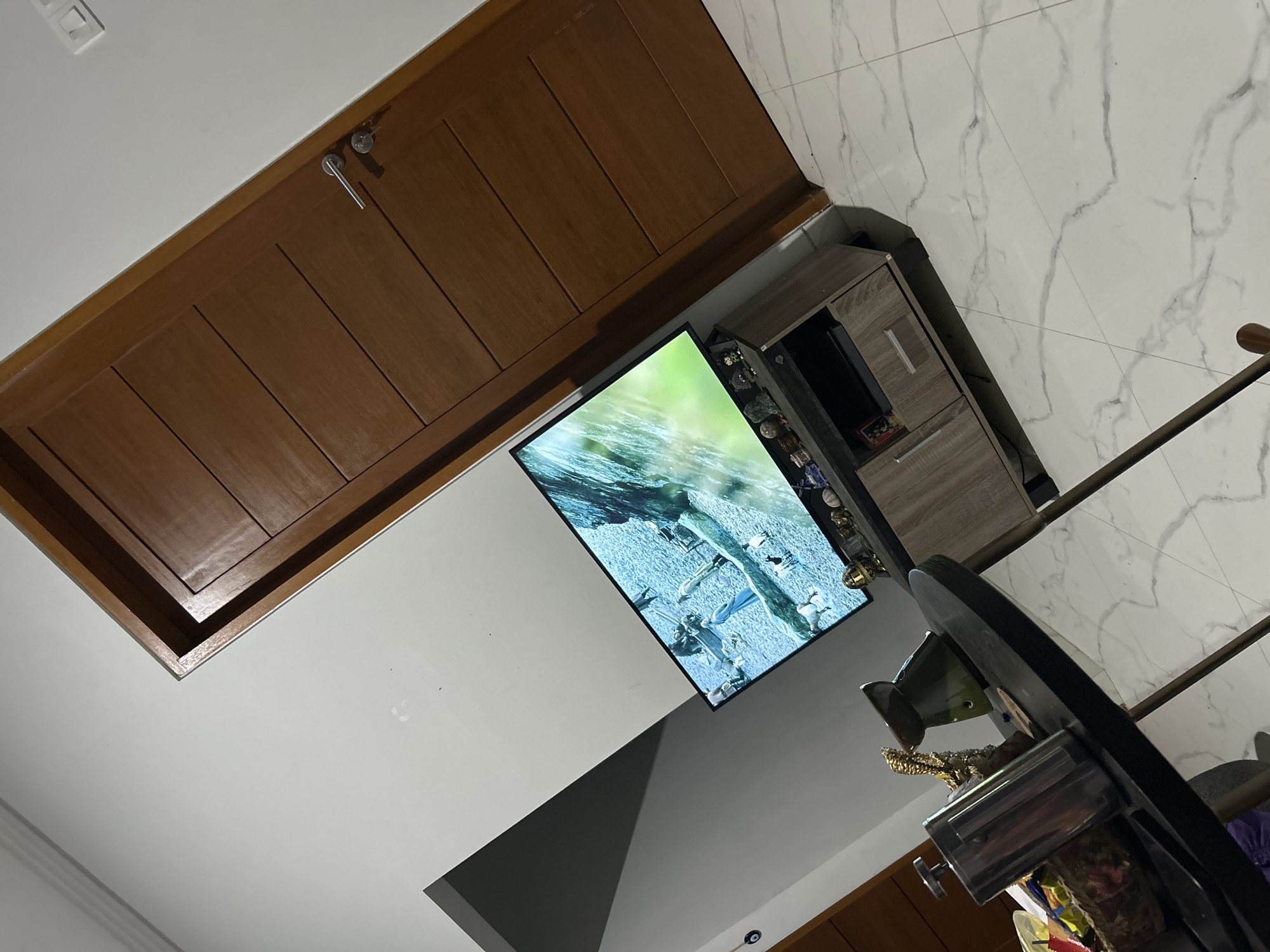 In Sukawati, Indonesia, a living room scene is captured, angled to show a television displaying a nature documentary or historical scene with animals and people. The TV is placed on a wooden entertainment center. To the right of the TV, a wooden door with horizontal paneling is visible. In the foreground, a dark, round table holds various objects, including a metal French press, decorative items, and a basket. The room features white walls and light-colored marble-patterned floor tiles. The lighting suggests it is daytime, with shadows cast by the objects in the room.