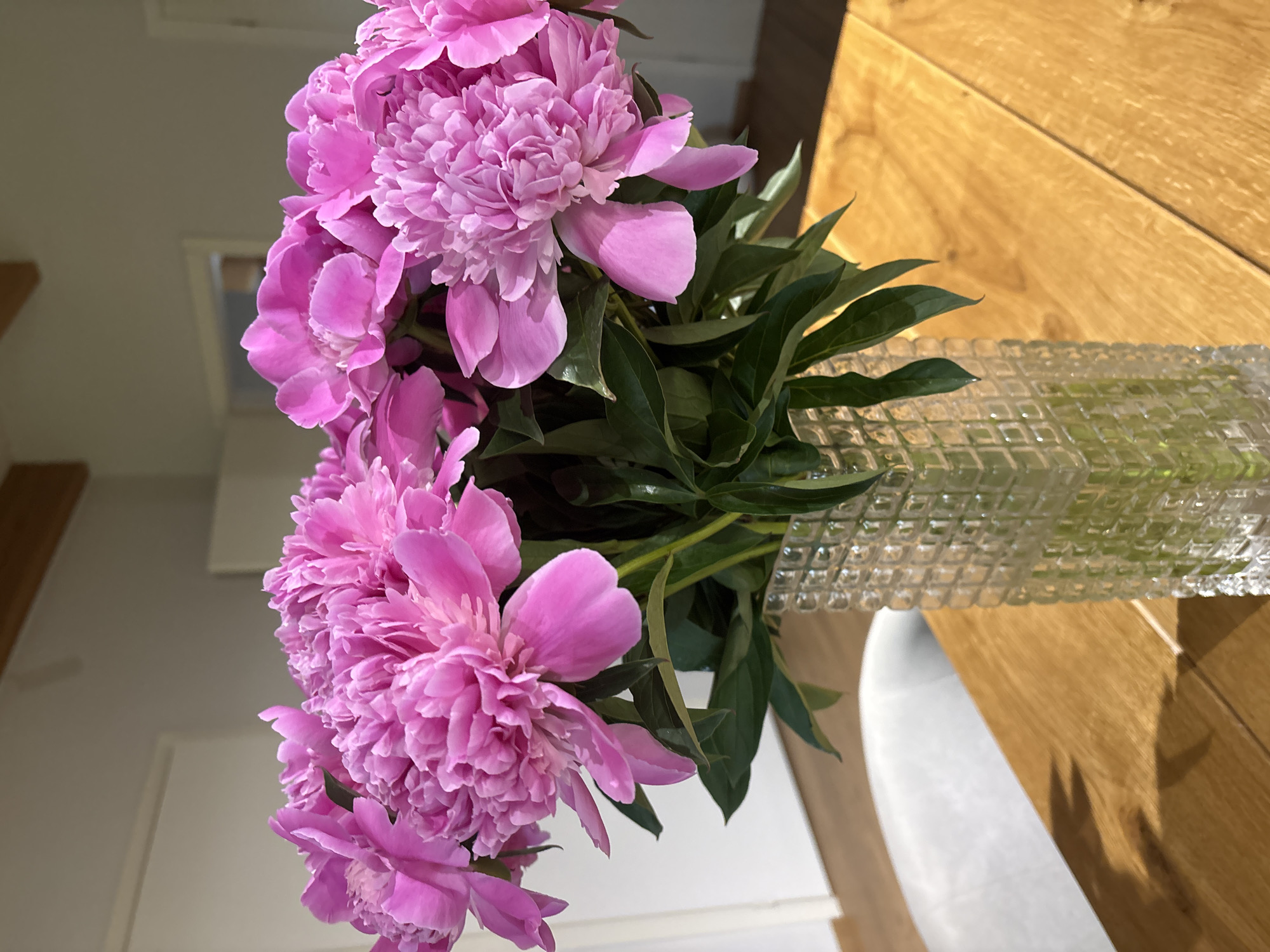 A bouquet of pink peonies in a clear, patterned vase sits on a wooden table. The table appears to be indoors, near a white wall and possibly a doorway. The specific location in Chaikovsky, Russia is not discernible from the image.