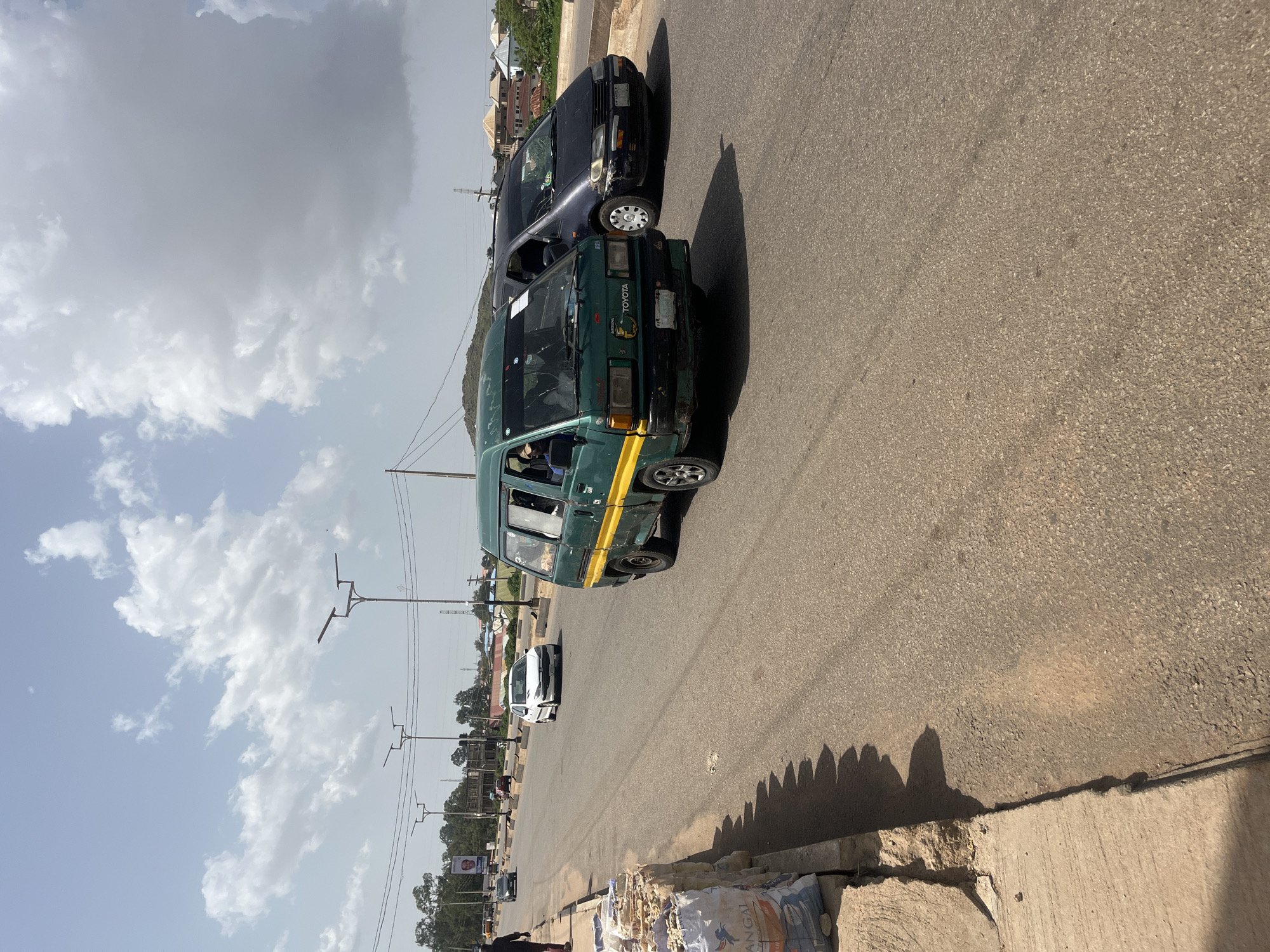 A street scene in Gura Top, Nigeria, shows a light green Toyota van with a yellow stripe driving on the right side of the road. To its right is a dark-colored minivan. In the background, other vehicles, buildings, and streetlights are visible under a cloudy sky. On the left side of the road, piles of bagged material are stacked near a curb.
