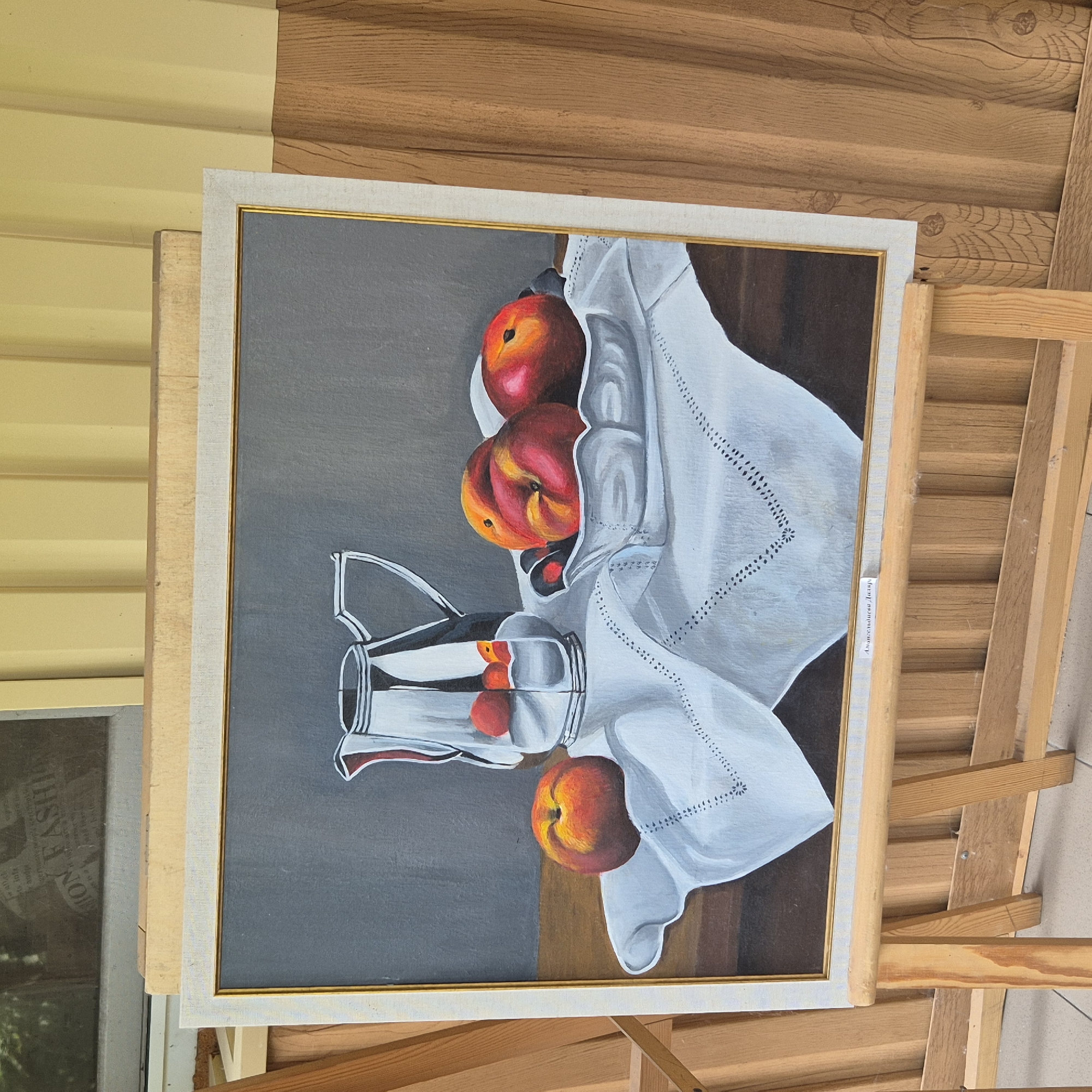 The image shows a still life painting of fruit and a pitcher, displayed on an easel outdoors. The painting features a silver pitcher, several peaches and dark berries in a decorative bowl, and a white tablecloth with embroidered details. The artwork is framed and leaning against a wooden structure, with a yellow corrugated wall and wooden planks visible in the background. There are no people present in the image. The scene appears to be in an outdoor setting, possibly a market or exhibition space, given the presence of easels and the general environment. There is no indication of Kokshetau, Kazakhstan.