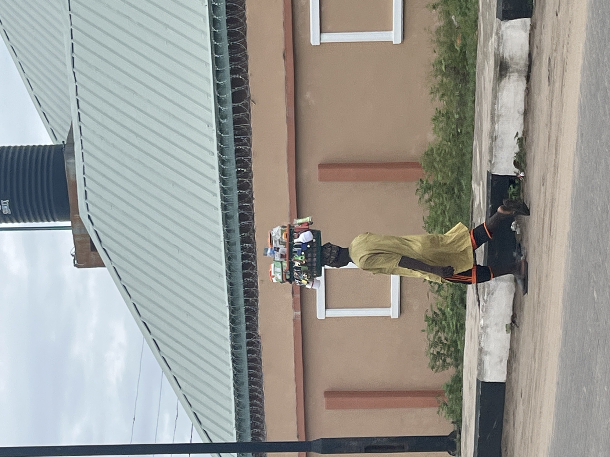 A street scene in Badagry, Nigeria, depicts an African man walking along a curb on a paved street. He carries a stack of goods balanced on his head. He is wearing a yellow long-sleeved shirt, black pants with orange stripes, and dark shoes. He is walking past a building with a light brown facade, windows framed in white, and green shrubbery along the base. Above, the building has a corrugated metal roof and a dark gray water tank. A security fence is installed along the roofline. In the foreground, a black metal pole runs horizontally across the lower portion of the frame. The sky is overcast.
