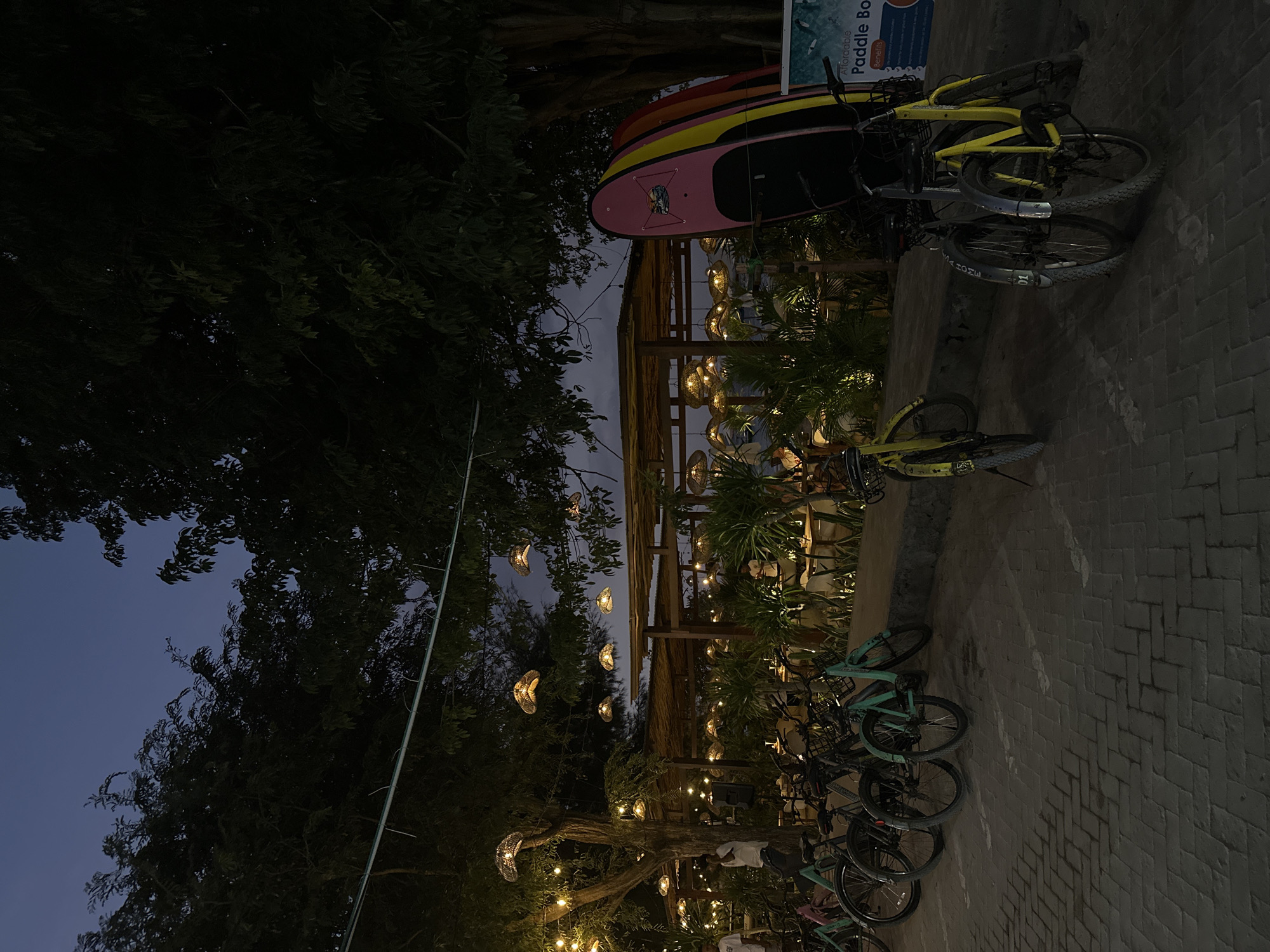 The image shows an outdoor scene in Unknown, Indonesia, at twilight or night. The location appears to be a recreation or leisure area, possibly near a beach. There are several bicycles parked in rows, some of which are yellow and others are teal. A stack of paddleboards rests against a structure. A partially visible sign mentions "Paddle Board." A wooden pergola-like structure is decorated with hanging lights, creating a warm glow. Trees with string lights are visible. Patrons are seated at tables. The ground is paved with brick.