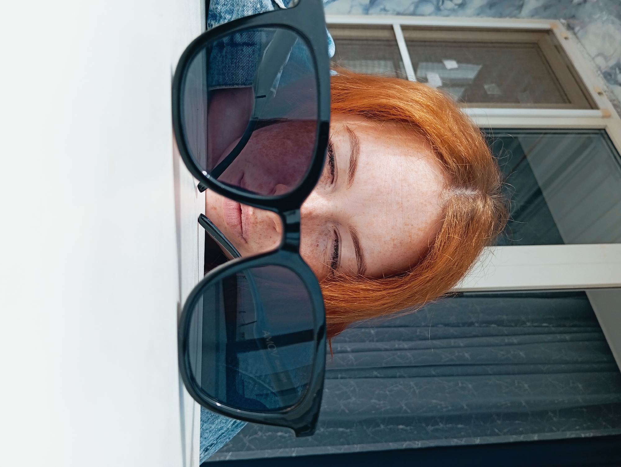 A portrait oriented close-up shot features a person with red hair and freckles, partially obscured by a pair of large, black-framed sunglasses held up to the camera. The person's eyes are closed, and their face is positioned between a white surface to the left and a window with a decorative blue-grey curtain in the background. A denim-jacketed shoulder is visible behind the sunglasses. The location is Yurgamysh, Russia.