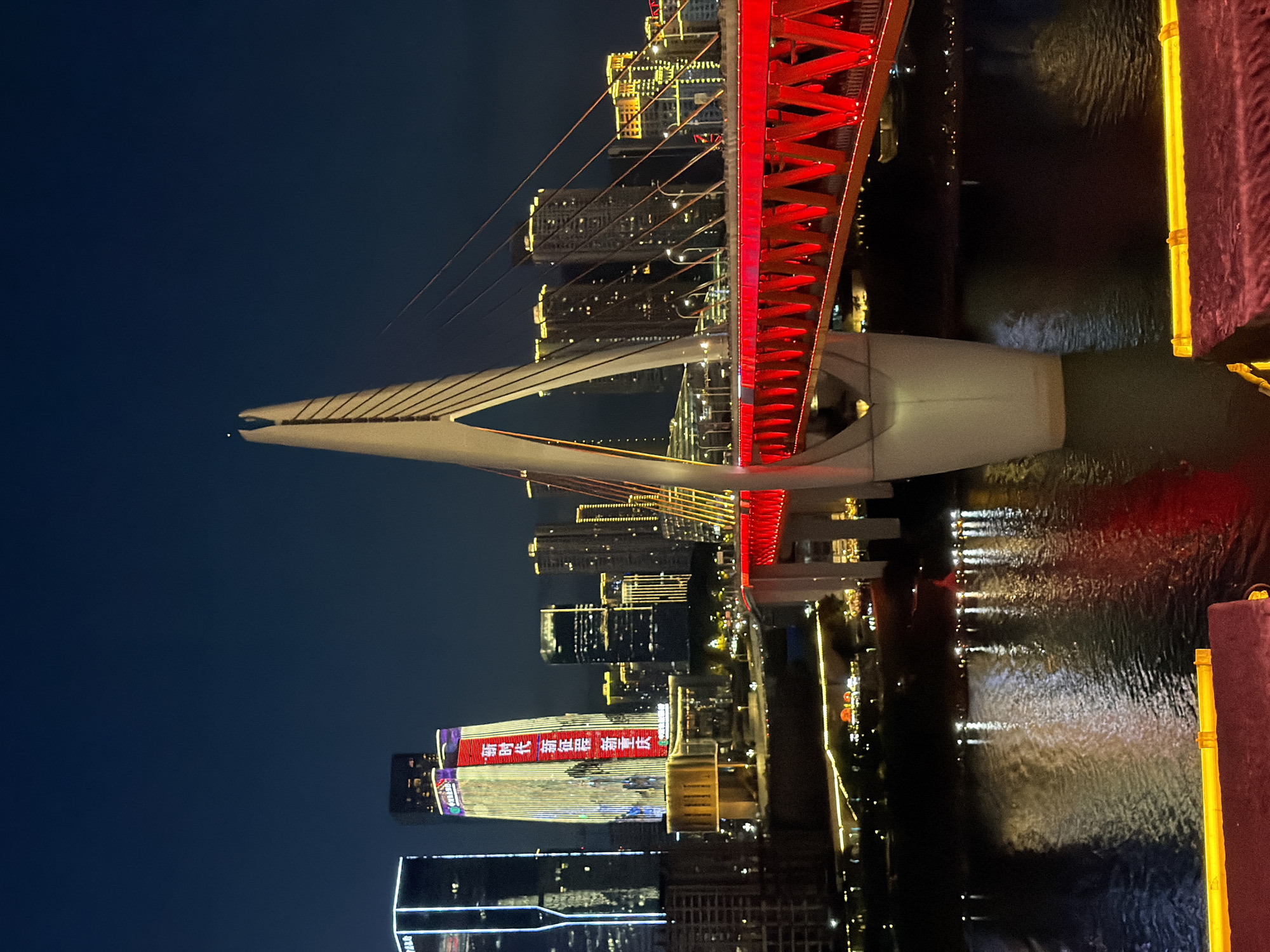 This is a night photograph of a modern suspension bridge illuminated in red, spanning a dark body of water. The bridge features a large, white pylon with cables supporting the roadway. The surrounding cityscape is visible in the background, with numerous tall buildings lit up with various colors of lights. One prominent skyscraper in the center has vertical red and yellow lights and a large red banner with white Chinese characters. The water reflects the lights from the bridge and buildings, creating a shimmering effect. The foreground shows the edges of what appear to be elevated seating areas with purple and yellow illuminated accents. The image is taken from a slightly elevated perspective, looking across the water towards the bridge and city skyline. The scene suggests an urban environment in Yuzhong District, China, likely at dusk or after dark, given the artificial illumination.