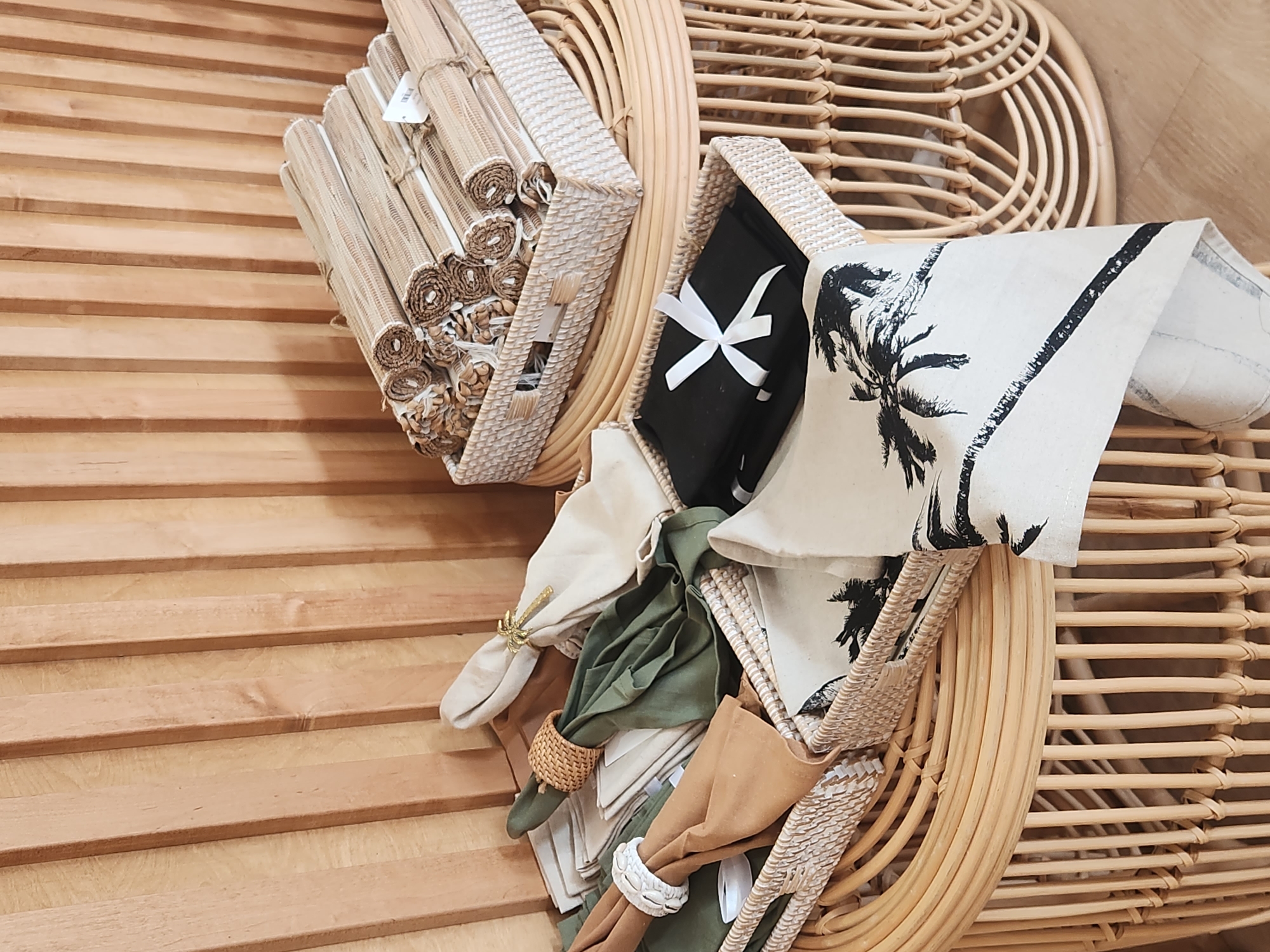 The image depicts a close-up, indoor shot of various table linens and placemats displayed on rattan furniture against a wood-paneled wall. The foreground features two woven baskets. The basket on the left contains folded napkins in shades of tan, sage green, and cream, some adorned with decorative napkin rings made of rattan and shells. Another basket, slightly further back, holds neatly stacked black folded fabric, tied with a white ribbon. On the right, a larger woven basket is filled with rolled-up placemats, appearing to be made of natural fibers, tied with twine. A white tag is visible on one of the placemat bundles. The rattan furniture, including a round table and baskets, has a light, natural wood finish. The background is a vertical wood-paneled wall with a warm, light brown tone. There are no people or animals visible in the image. The lighting is soft and diffused, suggesting an indoor setting. The image does not provide any specific information that would indicate its location within the city of Maitland, United States of America.
