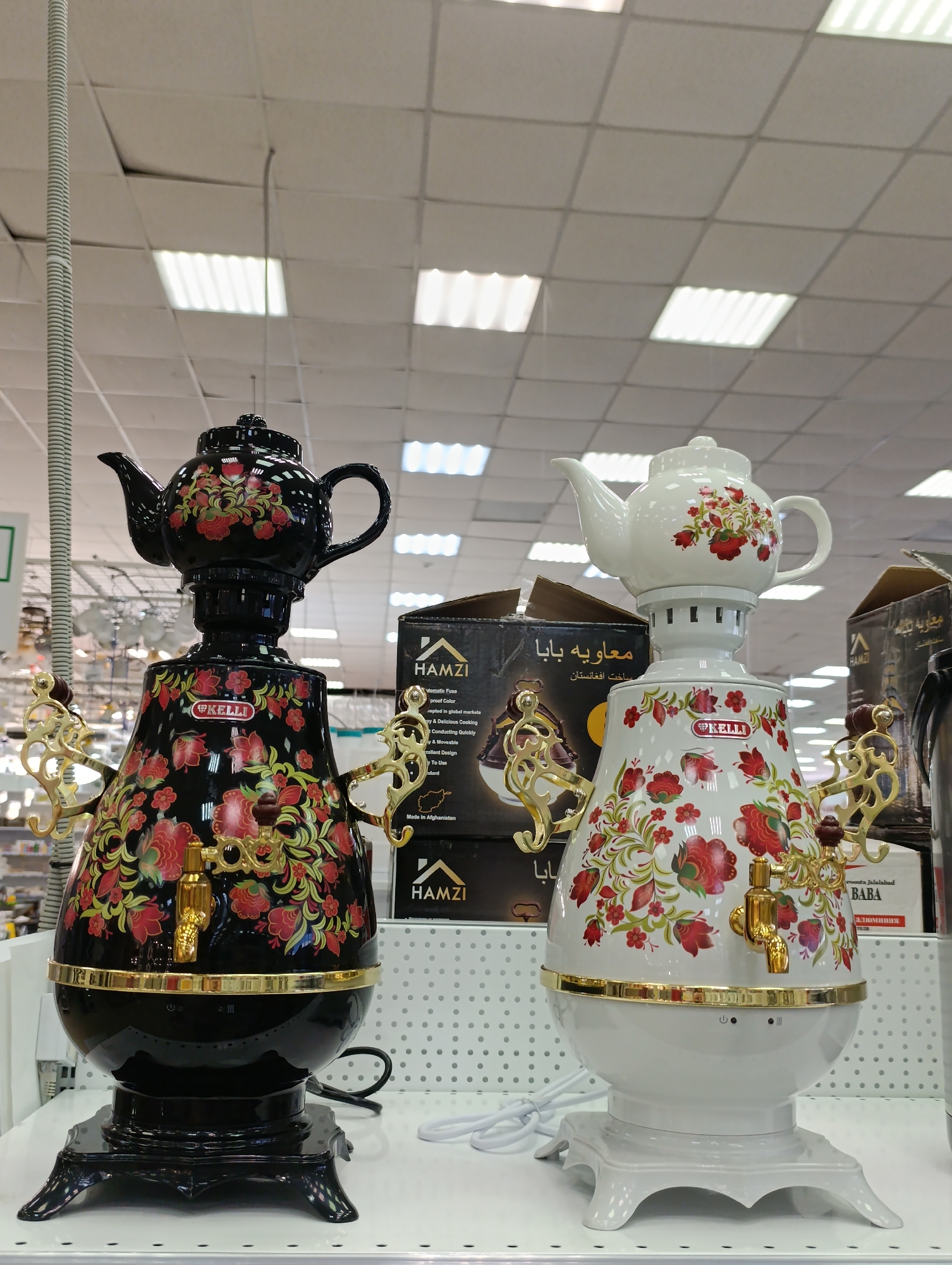 The image shows two decorative electric samovars, a type of heated metal container traditionally used to boil water, often for tea. The samovars are placed on a shelf under fluorescent lighting, likely in a retail setting.
One samovar is predominantly black with a floral pattern in red, green, and gold. The other samovar is white with a matching floral design. Both have gold-colored spigots and handles. Behind the samovars are boxes with Arabic and English text, including the brand "HAMZI," suggesting the origin or target market.
Based on text on one of the boxes, the products are "Made in Afghanistan". Additionally, the text says "roonata Jalalabad" - this is likely a misspelling of "roonta Jalalabad" which is a shop located in Chaikovsky, Russia.