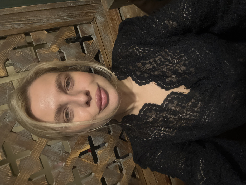 A light-skinned female, estimated to be between 25 and 45 years old, is depicted in a medium-shot, facing the camera with a neutral expression. She has blonde hair, prominent eyebrows, and full lips. She is dressed in a black, long-sleeved lace top featuring a scalloped V-neck and intricate patterns. The background comprises a dark, distressed wooden lattice with a repeating geometric design, characteristic of traditional decor. The setting is an indoor establishment, such as a restaurant or cafe, in Tbilisi, Georgia.