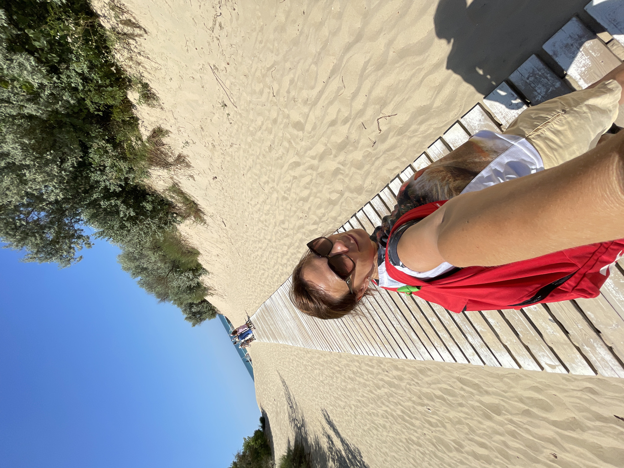 A woman is taking a selfie on a wooden boardwalk at a beach, with sand dunes and vegetation on her right and the sea and sky on her left. The boardwalk is made of weathered wooden planks and runs alongside a sandy beach. The woman is wearing sunglasses, a red backpack, and light-colored shorts. In the background, several people are walking along the boardwalk towards the sea. The sky is clear and blue, and the sun is shining brightly, casting shadows on the sand. The scene is likely located on one of the beaches of Anapa, Russia, known for its sandy shores and dune systems.