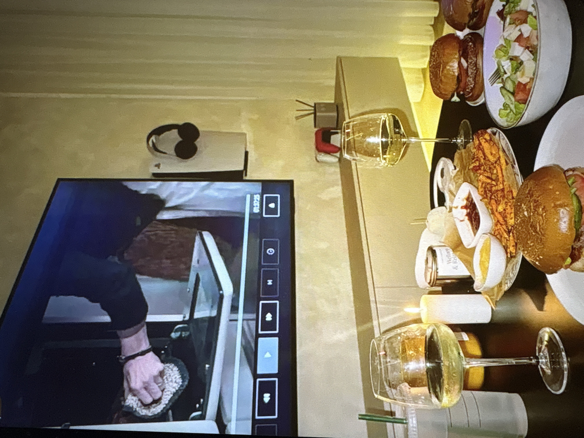 The image depicts a cozy home entertainment scene. In the foreground, a table is set with a meal, featuring burgers, fries, a salad, and drinks. Two wine glasses are filled with white wine, and a lit candle adds to the ambiance. The table is dark, contrasting with the lighter background.
The background features a large television screen displaying a cooking show. A person's hands are visible on the screen, cleaning a surface. On the wall next to the TV, a pair of headphones hangs on a white stand. A narrow shelf unit is mounted on the wall below the TV, holding a few decorative items, including a small red object and a container with reeds, possibly an air freshener. The wall is painted a pale yellow, and sheer curtains are visible on the right side of the frame.
The scene appears to be set in a domestic environment, likely a living room or dining area. There is no specific indication of the city of Pori, Finland within the image itself.