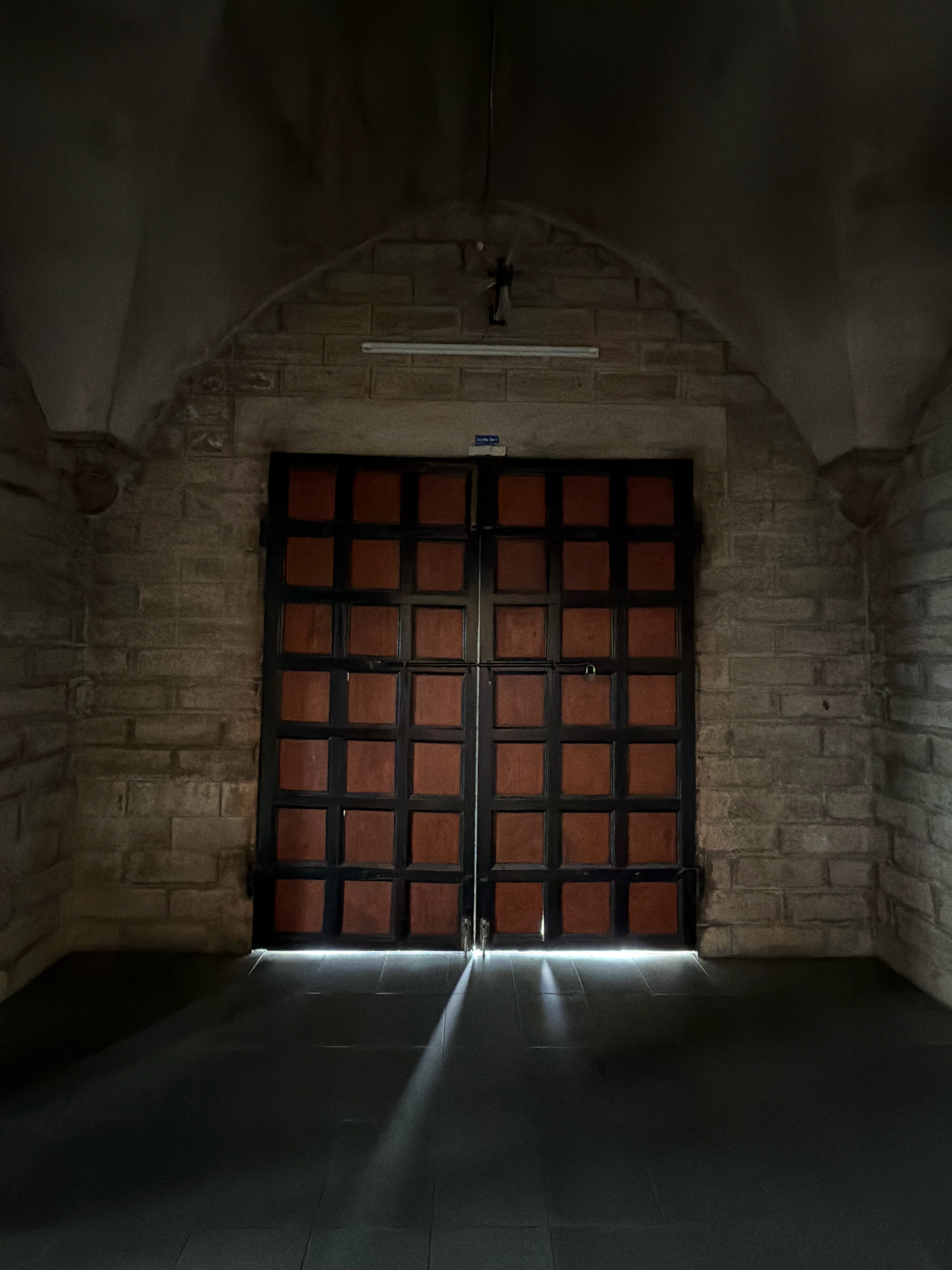 The image shows a pair of large, dark wooden doors with a grid of square panels, set within a stone archway in a dimly lit interior. Light streams from under the doors, casting long shadows on the dark tiled floor. The walls are constructed of light-colored, rough-hewn stone blocks. A fluorescent light fixture is mounted on the wall above the doors. The scene appears to be an entrance to an old building, possibly a historical site or religious structure. The specific location in Nha Trang, Vietnam is not evident from the image itself, but the architecture suggests a historical context.