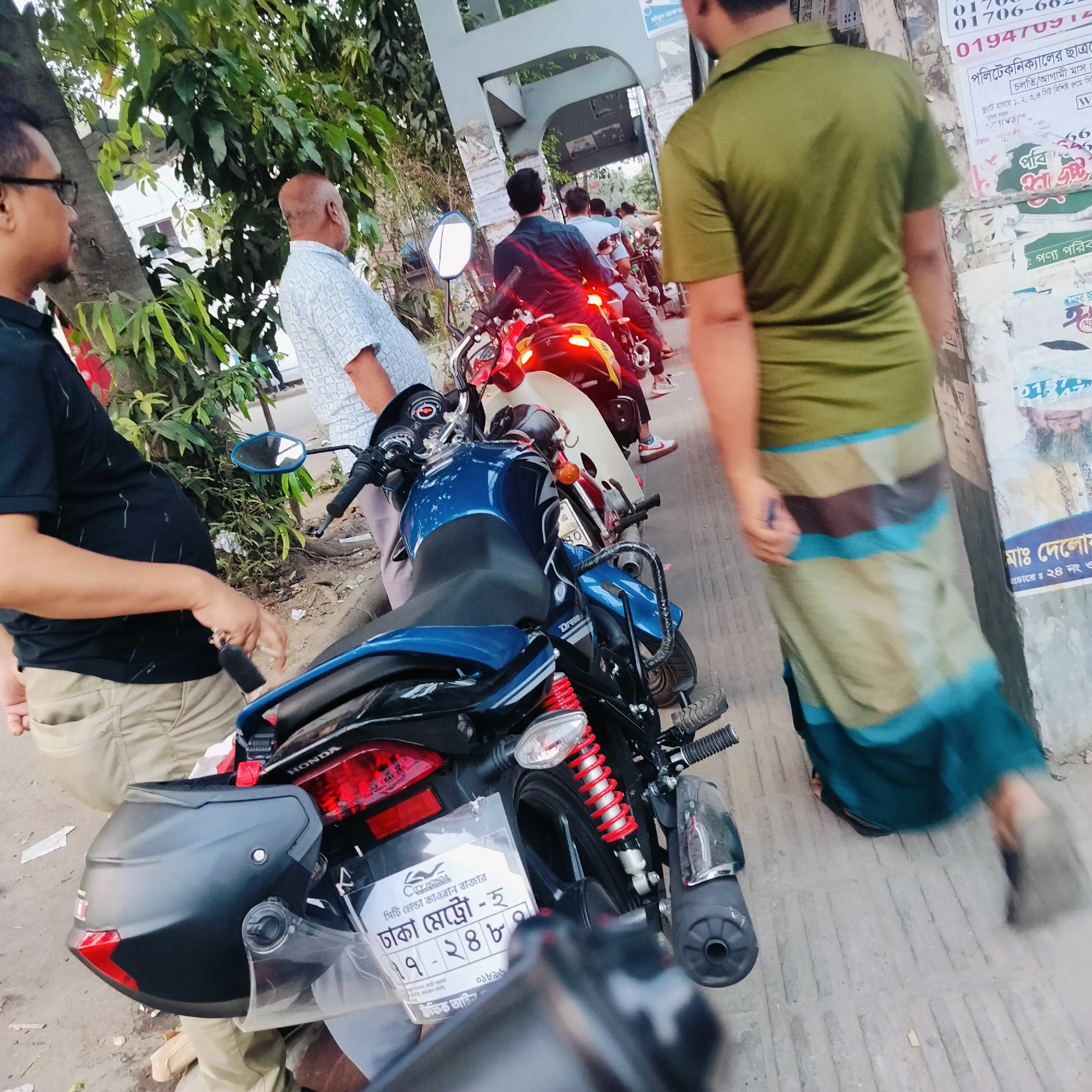 The image shows a street scene in Dhaka, Bangladesh. A line of motorbikes is parked on the side of the road, near a building. One of the motorbikes is a blue Honda with Dhaka metro registration number 17-2484. A helmet is strapped to the back of the motorcycle. A man is standing beside the motorbike with keys in his hand. Another man in a green shirt and lungi is walking past. Posters and advertisements are visible on a nearby wall, including phone numbers and mentioning "Polytechnic student". The poster displays the name "Ma: Deloar". A sign mentions the location as "City Honda Kawran Bazar". Other people are present, standing nearby. The environment appears to be urban, with trees along the street.