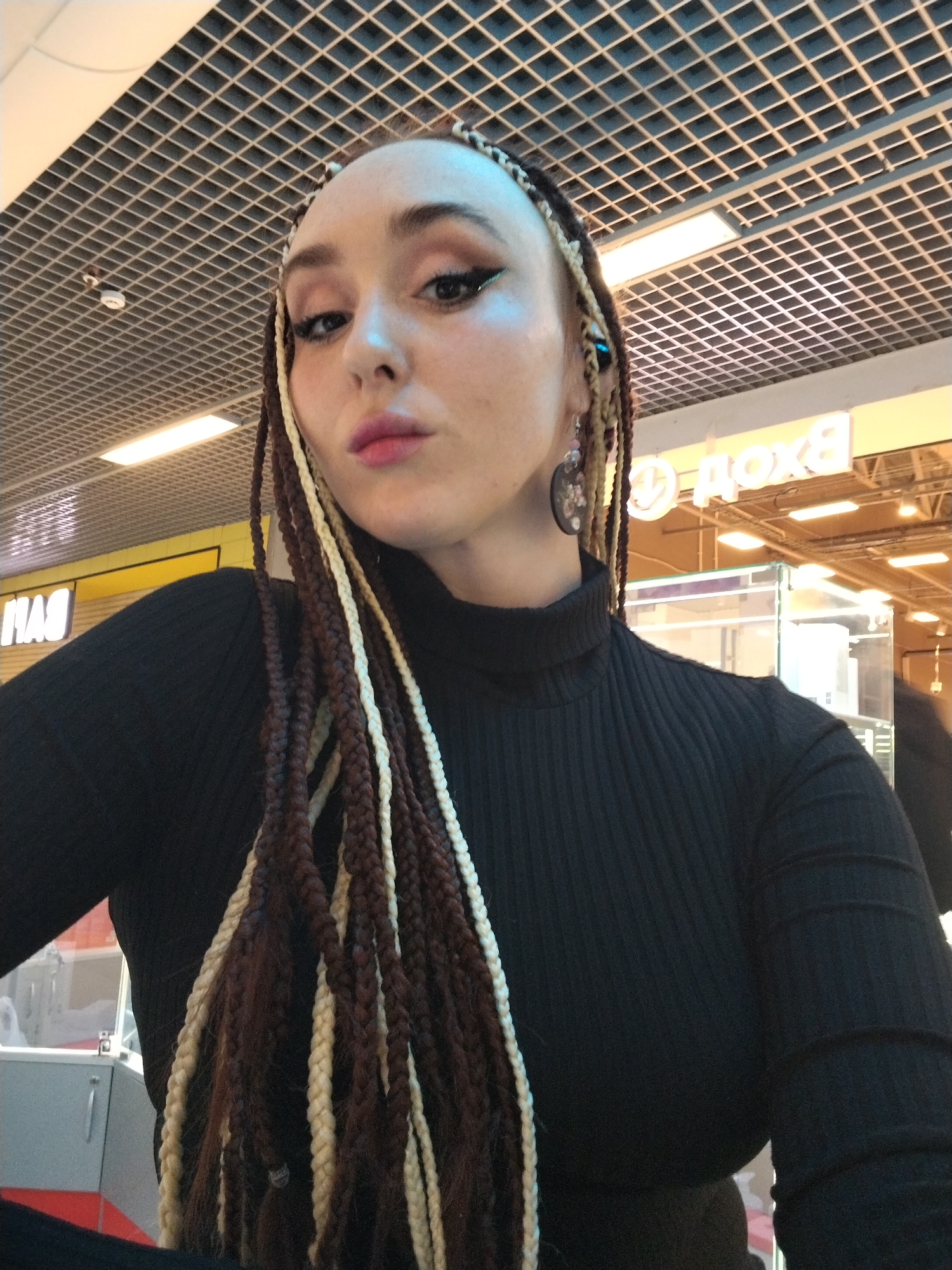 A woman is centered in the frame, appearing to be taking a selfie. She has light skin, makeup including winged eyeliner, and pink lipstick. Her hair is styled in long braids of brown and blonde. She is wearing a black turtleneck sweater.
The background is a commercial setting, likely a shopping mall. Above is a ceiling with a grid pattern and fluorescent lighting. In the midground, there are storefronts with signage in Cyrillic script, specifically the letters "ДОХОД", which could indicate a shop name or type of business. Display cases are visible within the stores.
The image was likely taken in Izhevsk, Russia, based on the Cyrillic text.