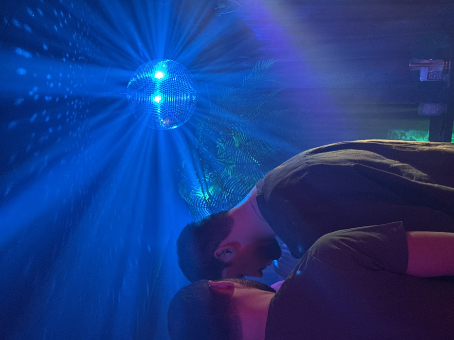 The scene depicts the interior of a nightclub or bar, presumed to be located in Yerevan, Armenia. The dominant illumination is a deep blue ambient light. In the upper right quadrant, a spherical mirrored disco ball is suspended, emitting numerous bright blue light rays that refract across the space and create scattered light reflections on the ceiling.
Two adult males are visible in the foreground, partially obscured. The individual on the far left presents their back to the viewer, wearing a dark short-sleeved shirt. To their right, a second individual is seen in left profile, facing slightly downward. This individual has short, dark hair and wears a dark-colored long-sleeved shirt with a visible metallic necklace or chain. No direct interaction between the two is discernible.
In the mid-ground, behind the individuals and beneath the disco ball, blurred green and blue illuminated botanical elements, resembling palm fronds, are present. The background to the lower right reveals a dark, horizontal surface on which at least two clear glass vessels are positioned, one appearing to contain a liquid or illuminated element.
