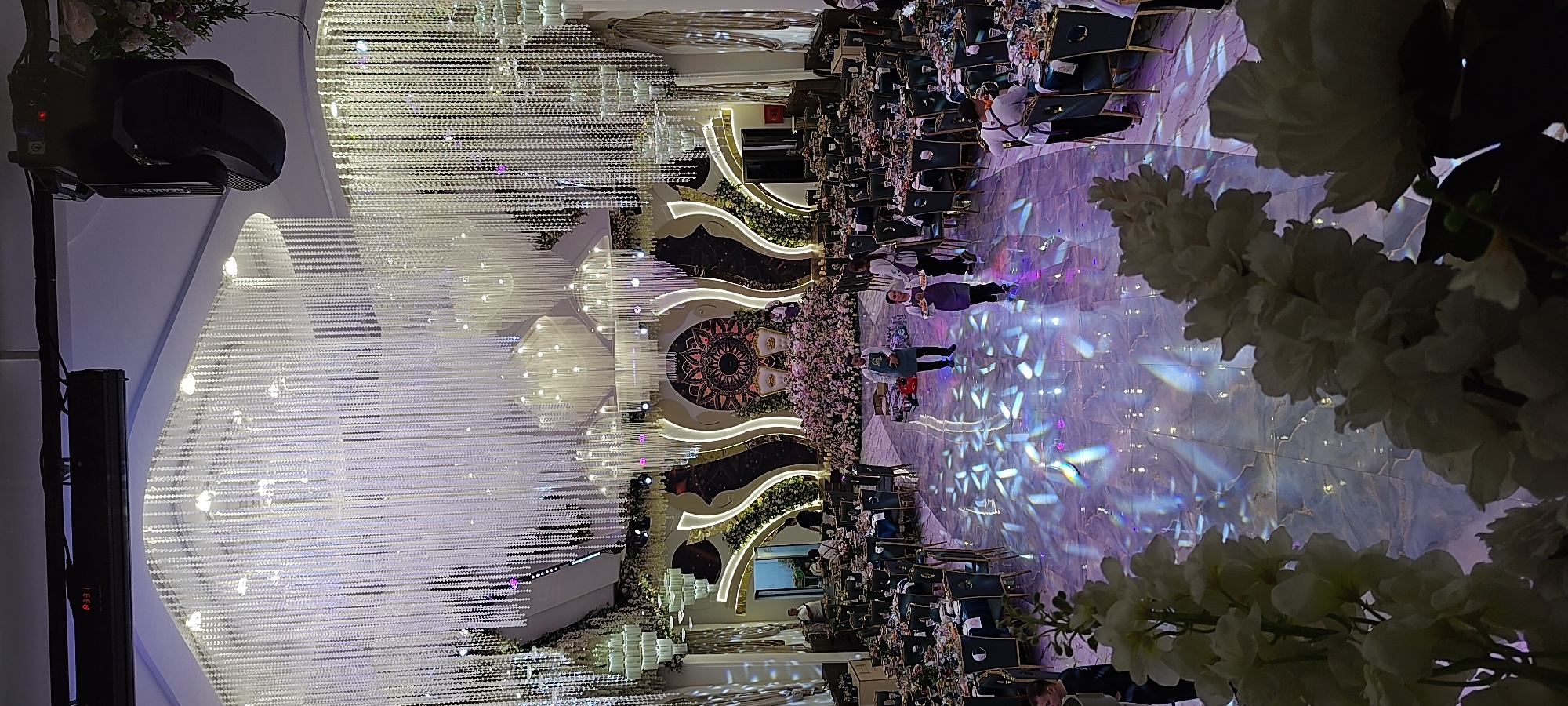 The image depicts the interior of a large, ornate event hall, likely a banquet hall or reception venue, in Khujand, Tajikistan. The hall is elaborately decorated with numerous crystal chandeliers hanging from the ceiling, creating a dazzling effect. The space is set up for an event, with many round tables draped in dark fabric and adorned with floral centerpieces. Chairs are arranged around the tables, indicating seating for guests.
In the background, a raised stage area is visible, featuring a large, circular floral arrangement behind two ornate chairs, suggesting it is a focal point for ceremonies or speeches. The walls of the hall are adorned with gold trim and decorative elements. The floor is highly polished, reflecting the overhead lights and creating a kaleidoscopic pattern of light and color across the surface.
Several people are present in the hall, engaged in various activities. Some individuals appear to be staff members, possibly waiters or event organizers, as indicated by their attire, such as aprons. One woman is seen carrying a tray of what appears to be food. Other individuals are mingling or attending to preparations.
The overall ambiance is one of luxury and festivity, characteristic of a high-end event venue. The lighting, decorations, and table settings all contribute to a grand and sophisticated atmosphere. The presence of floral arrangements, both on the tables and the stage, further enhances the celebratory feel. The detailed decorations and expansive layout suggest a venue designed for significant social gatherings and celebrations.