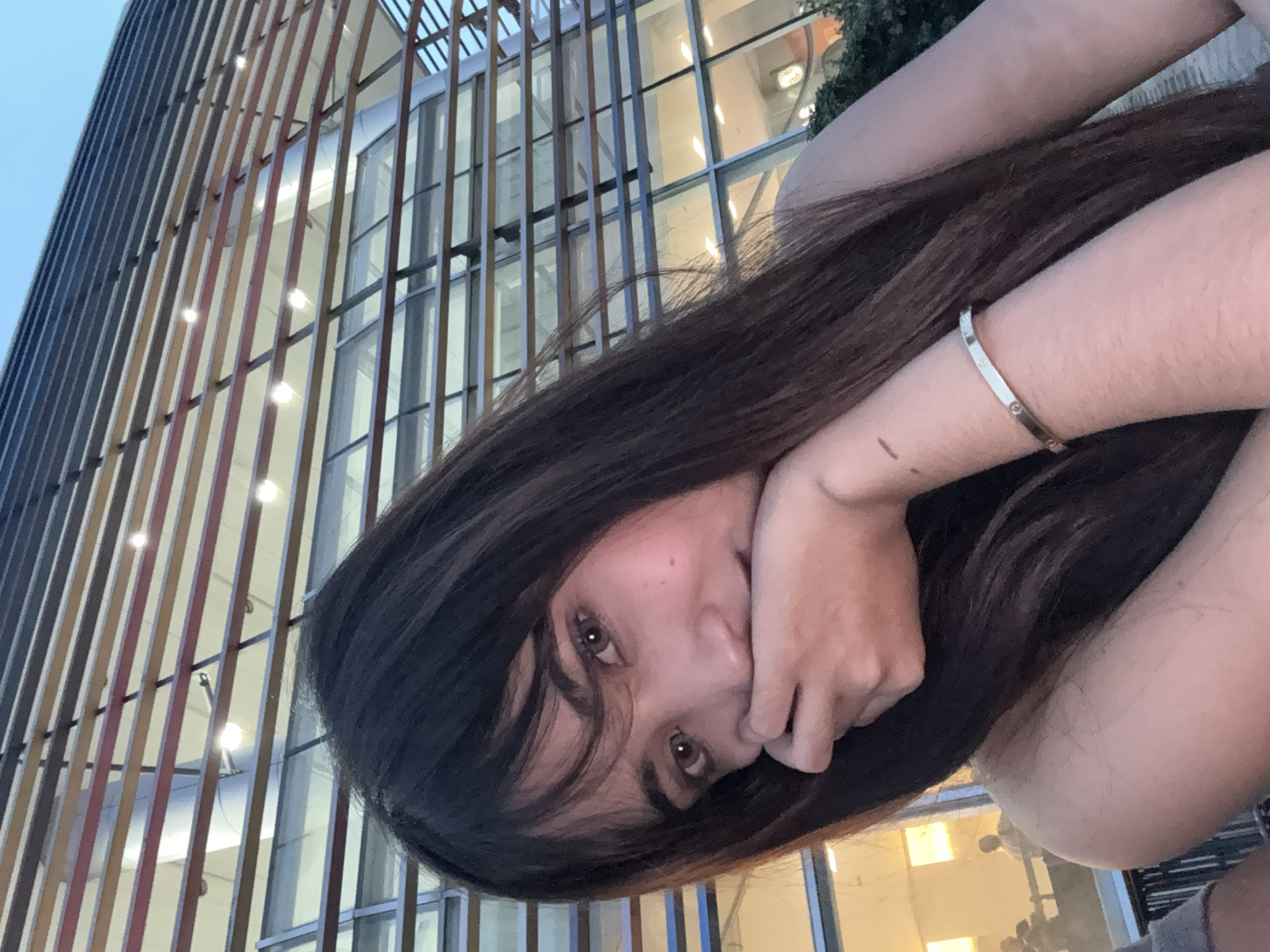 A young woman with long dark hair is facing the camera. She is partially covering her mouth with her hand. A silver bracelet is on her wrist. The background includes a building with vertical slats. This building appears to be located in Davao City, Philippines.