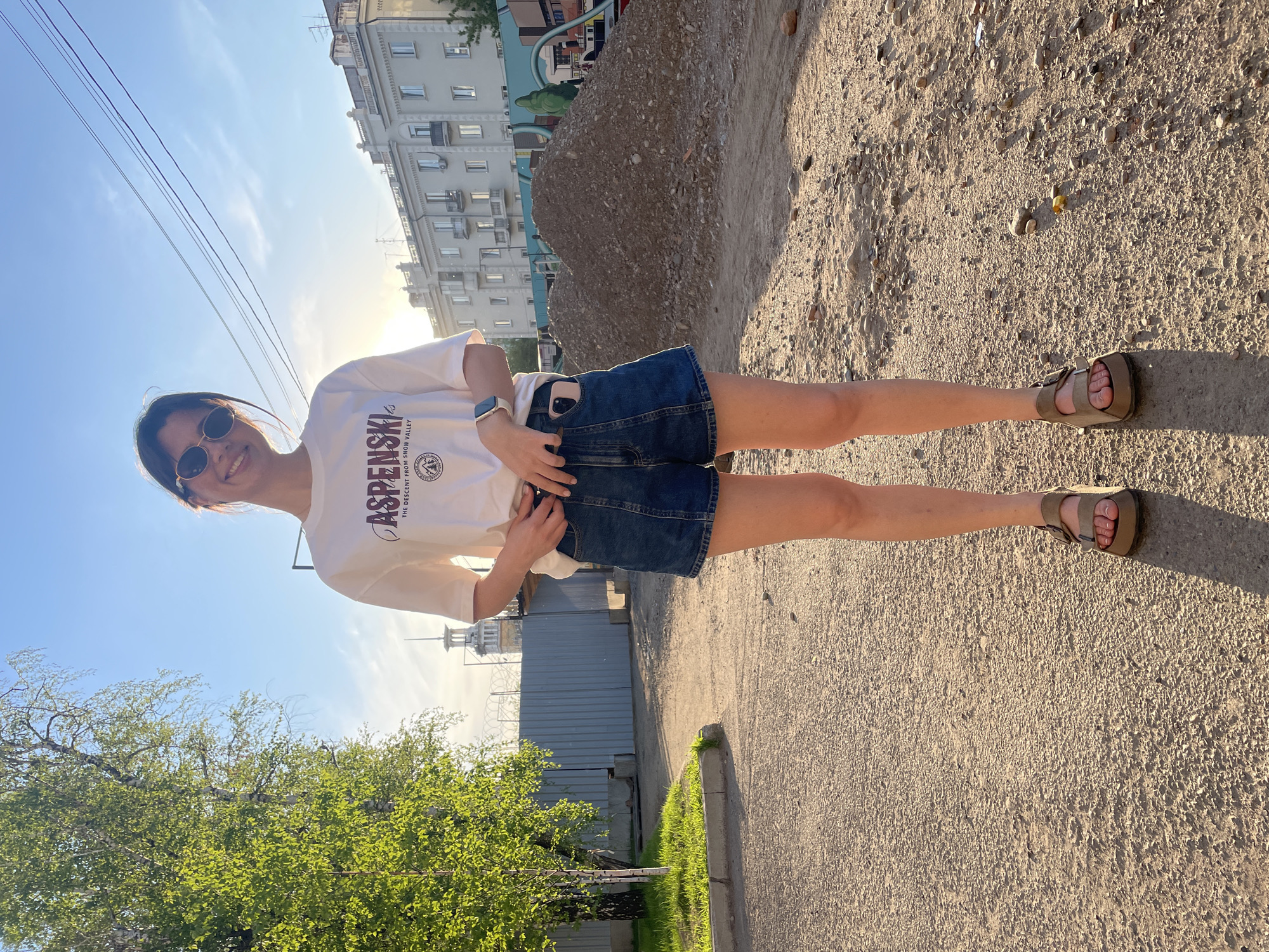 The image captures a young woman standing outdoors, smiling directly at the camera. She is wearing a white short-sleeved t-shirt with the text "ASPENSKI" prominently displayed in maroon, along with "THE DESCENT FROM SNOW VALLEY" and a circular logo beneath. She pairs this with blue denim shorts, and light brown flat sandals. Dark, round sunglasses are on her face, and a light-colored smartwatch is visible on her left wrist. A white phone is tucked into the back pocket of her shorts. She stands with her hands near her waist, striking a casual pose.
The setting appears to be an urban or semi-urban environment in Krasnoyarsk, Russia, under a clear blue sky. The sunlight is warm and bright, suggesting late afternoon or early evening, with long shadows visible on the ground. In the background, there are multi-story residential or commercial buildings. Power lines stretch across the upper part of the frame. To her left, lush green foliage of a tree is visible, while to her right and behind her, there are large piles of earth or rubble, indicating either a construction site or an unpaved area. The ground beneath her is a mix of dirt, gravel, and rough pavement. A section of a grey corrugated metal fence is also visible in the mid-ground. The overall impression is a cheerful, sunny day.