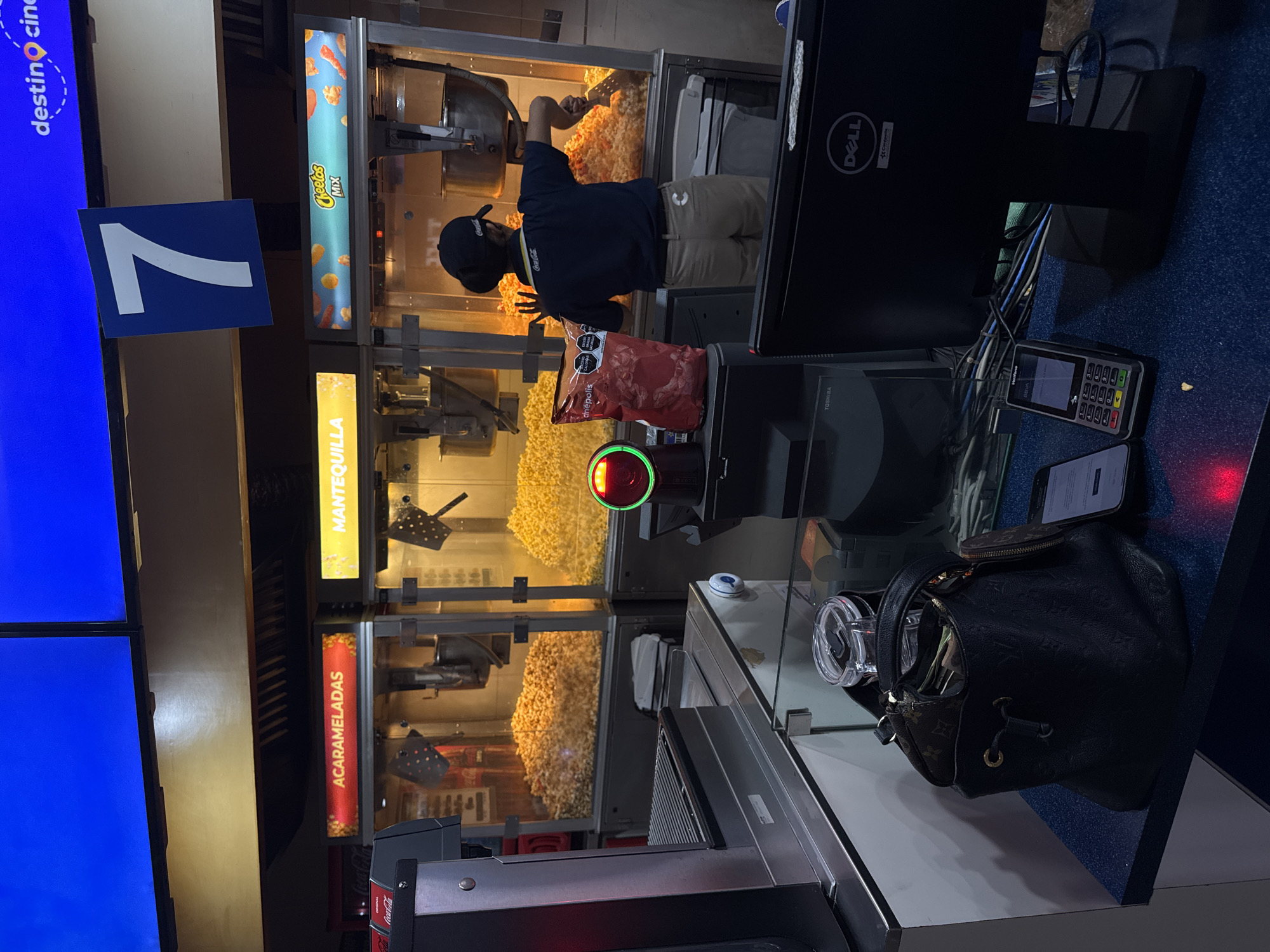 Here's a detailed description of the image:
**Location:** This scene appears to be inside a Cinepolis movie theatre in the city of Puebla, Mexico, based on the text visible on the screens.
**People:** A person is shown working behind the concession counter, wearing a dark Coca-Cola branded baseball cap, a dark t-shirt with Coca-Cola branding, and light brown pants. The individual appears to be preparing popcorn using a shovel.
**Subjects/Objects:**
* Popcorn machines: Several popcorn machines are visible, labeled with flavors such as "Mantequilla" (Butter), and "Acarameladas" (Caramelized). One is labelled "Cheetos Mix."
* Popcorn: The machines are filled with popcorn.
* Snack bags: A bag of Cinepolis-branded snacks is on the counter.
* Point of Sale (POS) System: There's a Dell monitor, Toshiba terminal, a VeriFone credit card terminal, and a smartphone on the counter, forming the POS system.
* Handbag: A black handbag with visible brown branding (likely Louis Vuitton) is sitting on the counter.
* Signage: A blue sign with a white number "7" is visible.
* Coca Cola advertisement: A sign that states "Coca Cola" in red and white.
**Actions:** The employee is in the process of preparing popcorn.
**Environment:** The scene is a brightly lit concession stand, possibly in a movie theatre.