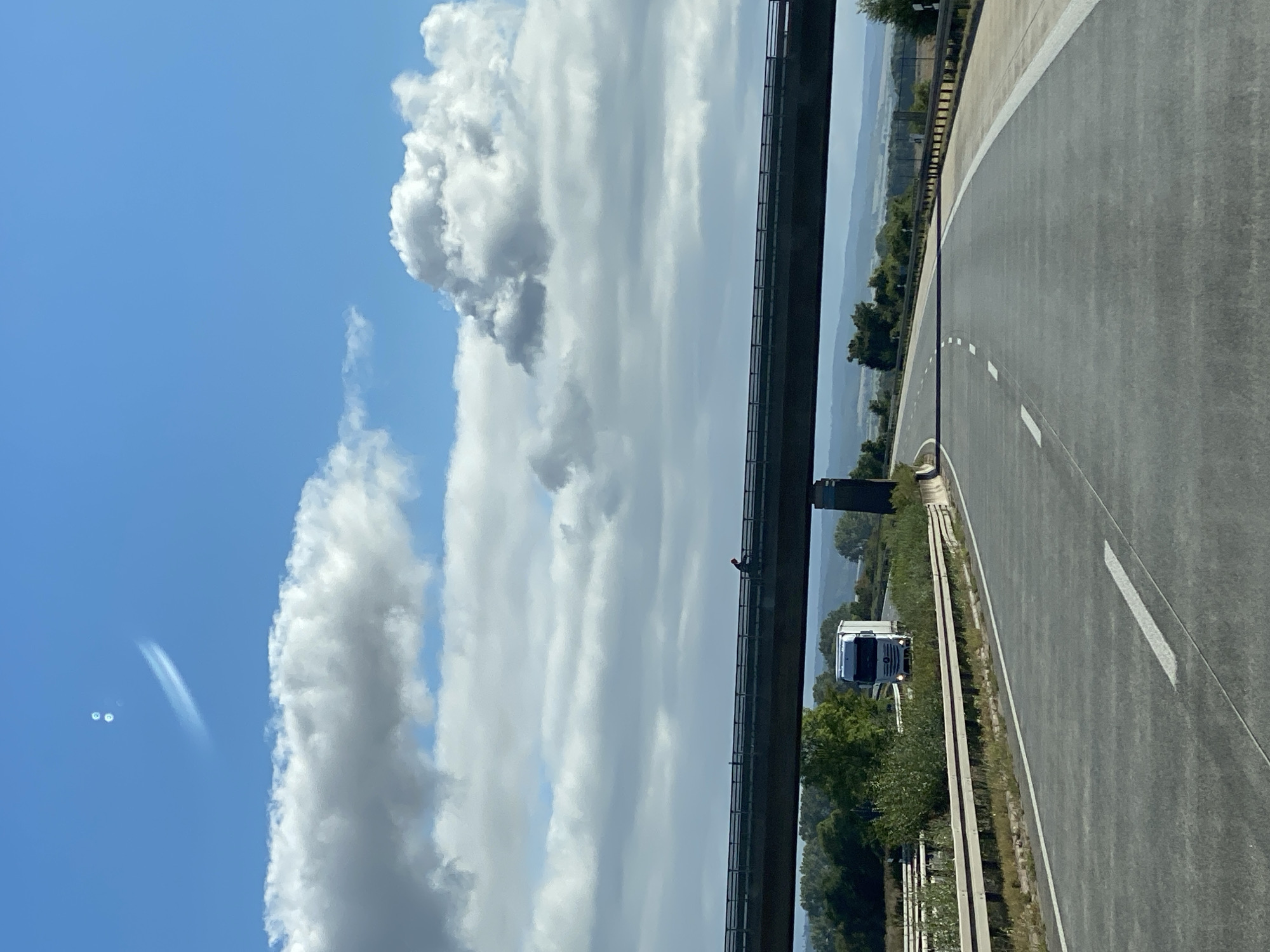 A highway overpass looms over an empty highway under a bright blue sky with scattered clouds. A single figure, wearing a red garment, is visible walking on the overpass. Below, a white truck is driving on the highway. The scene appears to be a rural area near Nordhausen, Germany. The image also features a peculiar atmospheric phenomenon in the sky, with two circular bright spots and a wispy trail, possibly contrails from an aircraft. The overall impression is one of a quiet, ordinary day on a German autobahn, with an unusual sky.