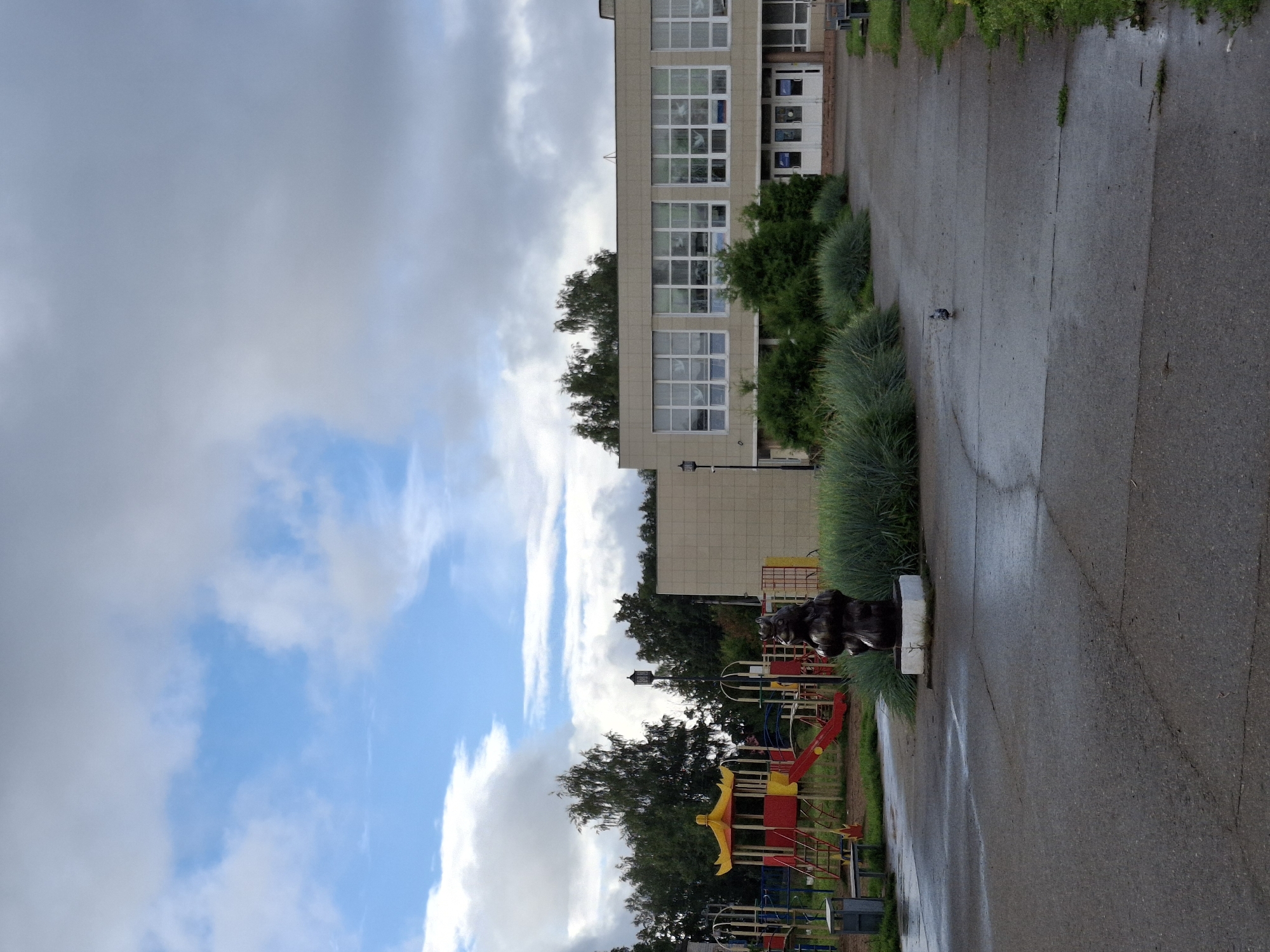 The image depicts a view of a schoolyard or public space in Elovo, Russia. A light-colored, modern building with large windows forms the backdrop. In the foreground, a wet asphalt path leads towards the building. To the left, a colorful children's playground with slides and climbing structures is visible, partially obscured by trees. A dark, abstract sculpture stands on a concrete base near the playground, surrounded by lush green bushes. Further down the path, a single pigeon is walking. The sky is partly cloudy with patches of blue. The overall impression is one of a functional outdoor space, likely a school or community center, during overcast weather.
