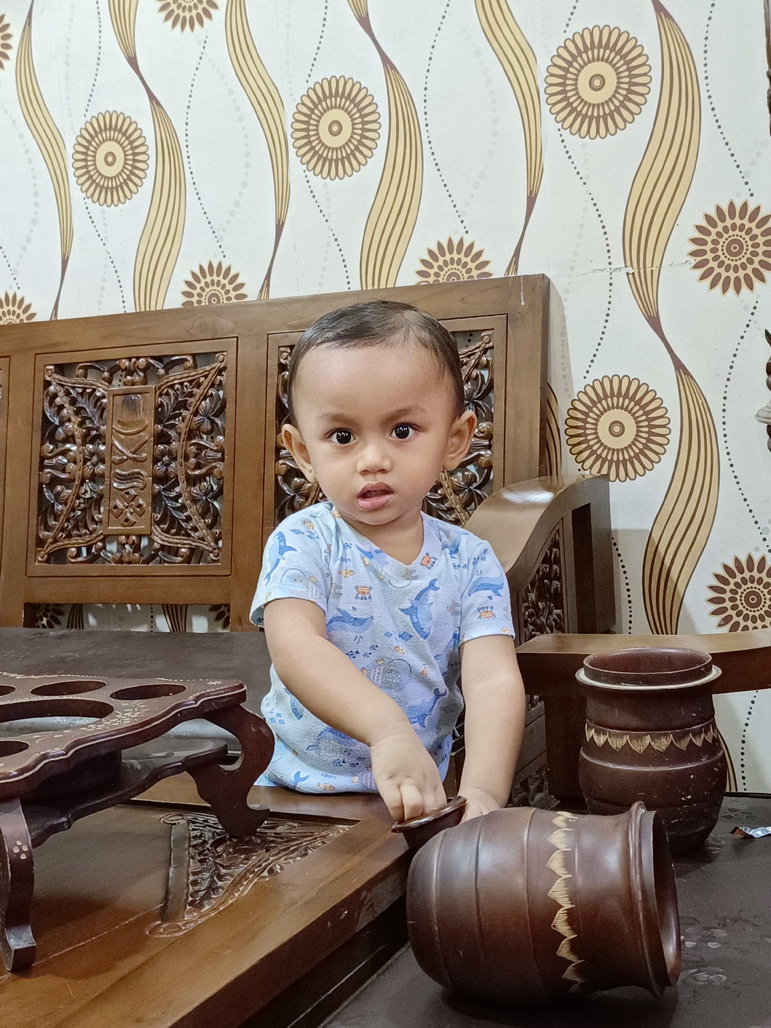 A young child, appearing to be between 1 and 2 years old, sits at a dark wooden table in front of a wooden furniture piece with intricate carvings. The child is wearing a light blue t-shirt with a pattern of blue whales and other sea creatures. The child's gaze is directed towards the camera with a neutral expression. The background features wallpaper with a repeating pattern of stylized floral motifs in yellow and brown on a white base. On the table, to the child's right, are two dark brown, lidded ceramic vessels with decorative patterns. To the child's left, on the table, is a dark brown wooden stand with circular indentations. The scene appears to be indoors, possibly in a home, and the overall setting is consistent with a residential interior in Blang Cut, Indonesia.