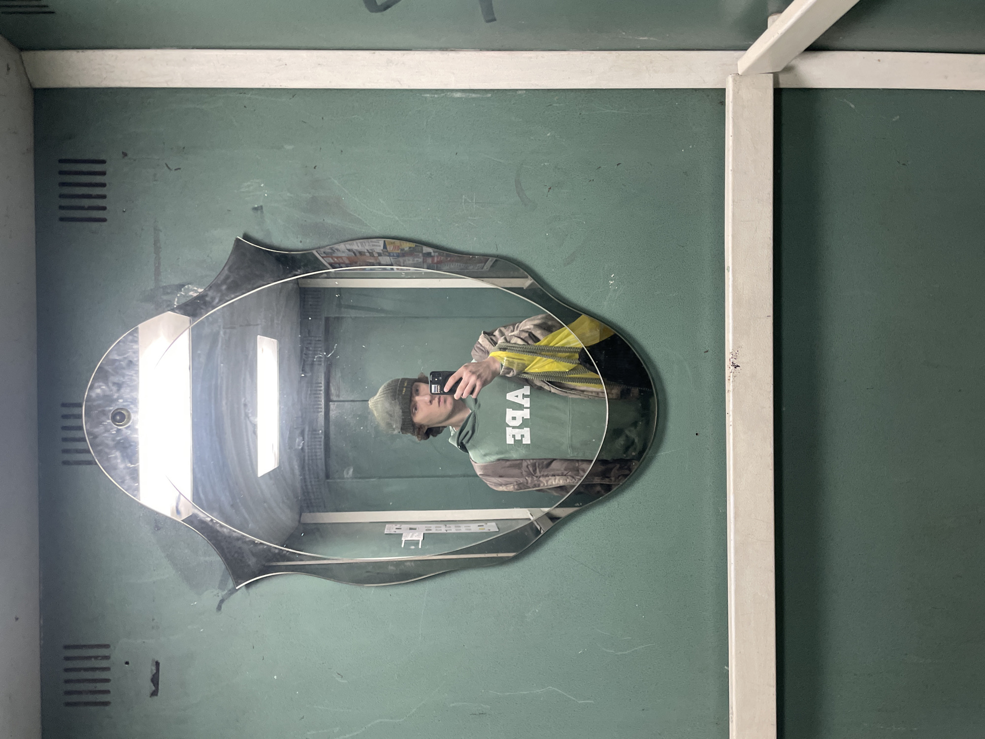 A person is reflected in an oval, decorative mirror attached to a faded green wall. The person is wearing a green beanie and a green hoodie with white lettering. The person is holding a mobile phone and taking a picture of themselves. The mirror is mounted on the wall of what appears to be an elevator car. The overall environment seems utilitarian and slightly worn. It is plausible that the location is Krasnoyarsk, Russia, based on the visual characteristics of the image, such as the aesthetic of the elevator and wall, which are typical of many older Russian buildings.