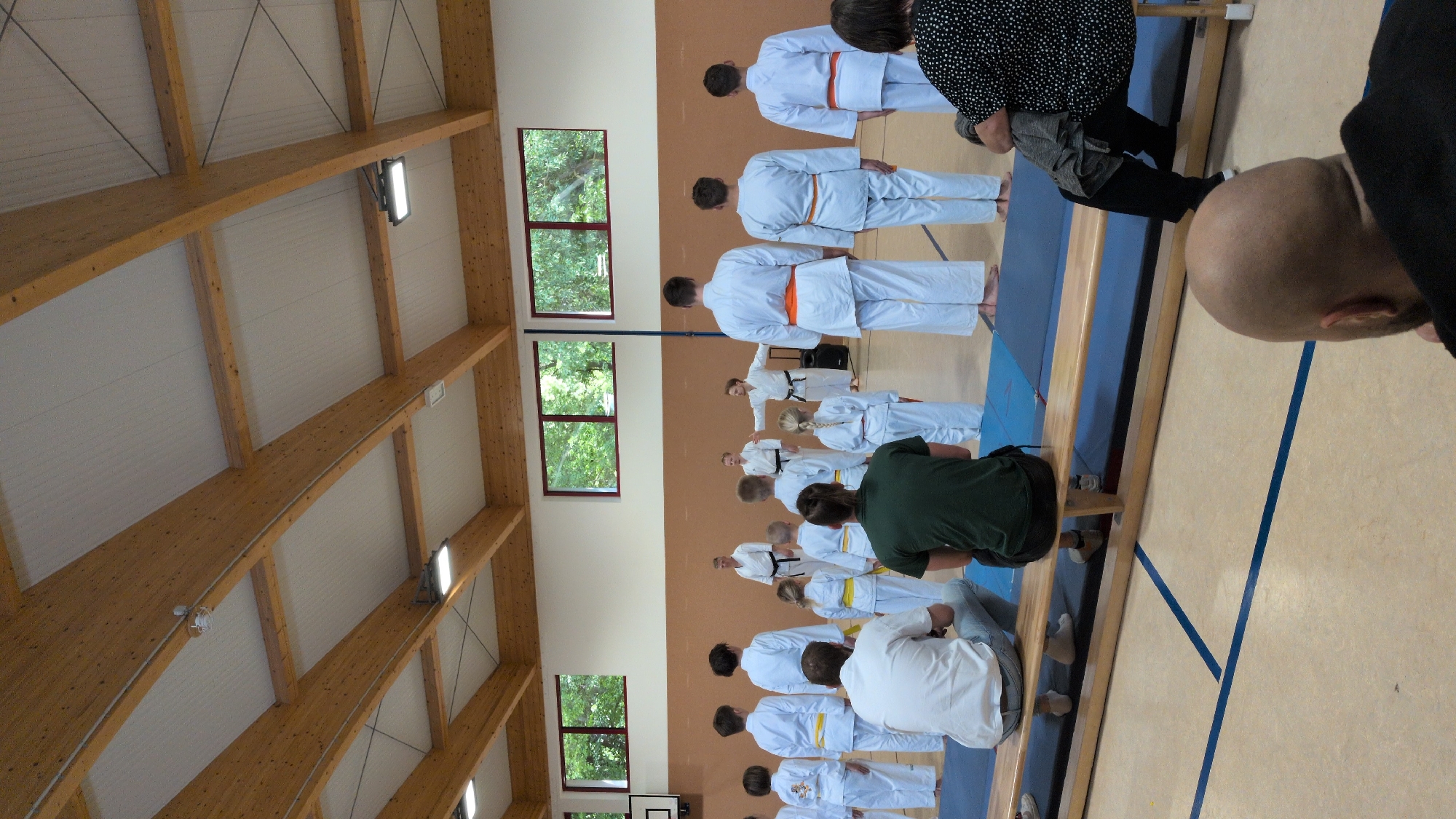 A group of people are standing in a gymnasium in Neudietendorf, Germany, facing a man in a white karate uniform with an orange belt, who is gesturing with his hands. Several children and adults, also in white karate uniforms, are lined up. Some have yellow belts, while others have no belts visible. A few adults are seated on a wooden bench in the foreground, facing away from the camera. The gymnasium has a wooden ceiling with exposed beams and lighting fixtures. Windows with trees visible outside are present on the far wall. The floor is a light color with blue lines.