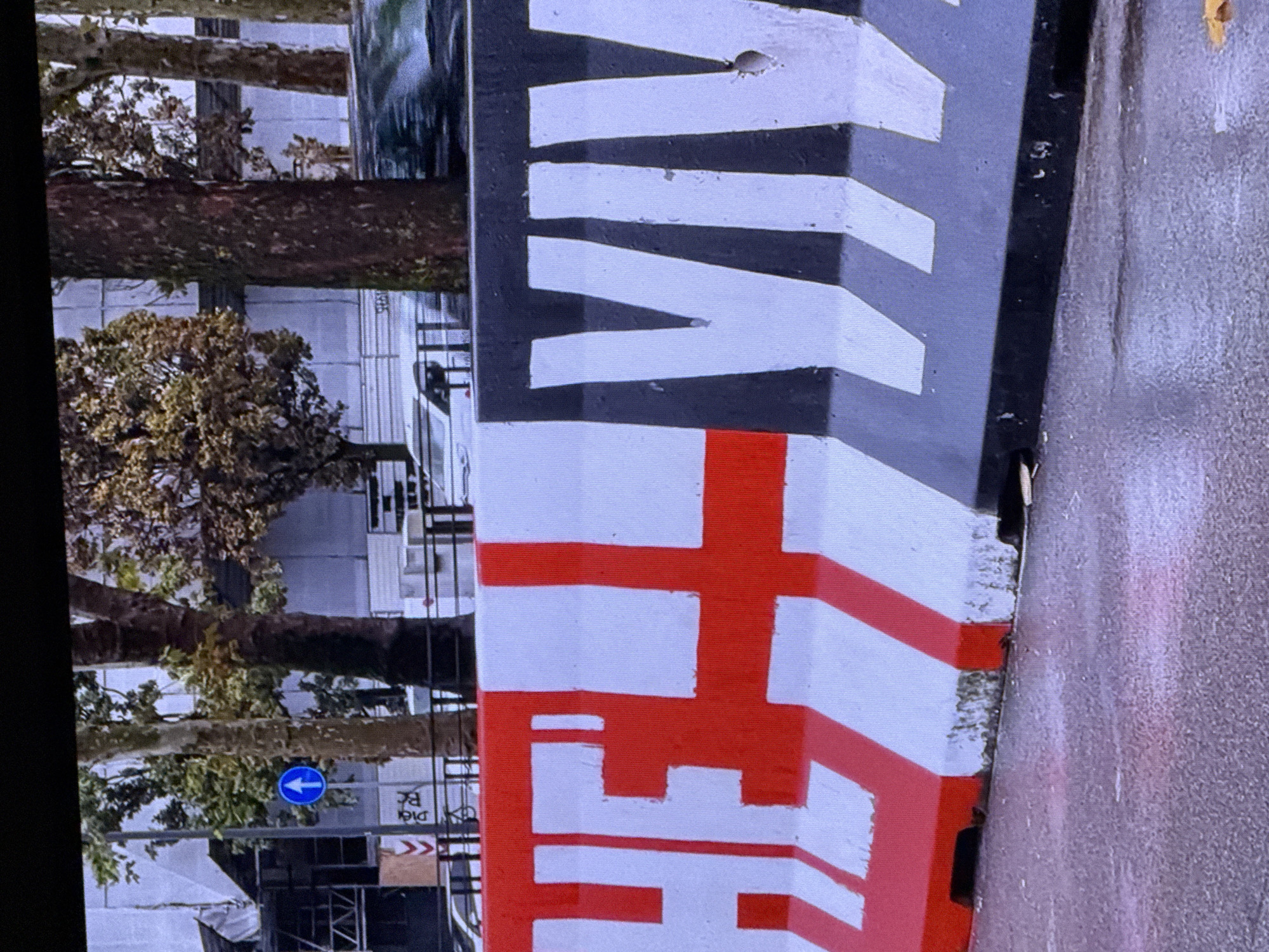 The image shows a close-up of concrete barriers painted with large white letters on red and black backgrounds. On the left, the letters "HELL" are visible in white on a red background, with white vertical lines also painted on the barrier. On the right, the letters "VIV" are visible in white on a black background, with white diagonal lines. The barriers are placed on a wet asphalt surface, suggesting it may have recently rained. In the background, out of focus, are trees with sparse autumn foliage, buildings, and a road with a car in motion. There is also a blue circular traffic sign with a white upward arrow visible in the upper left portion of the image. The scene appears to be an urban environment. There is no indication that this image is from Dubai, United Arab Emirates.