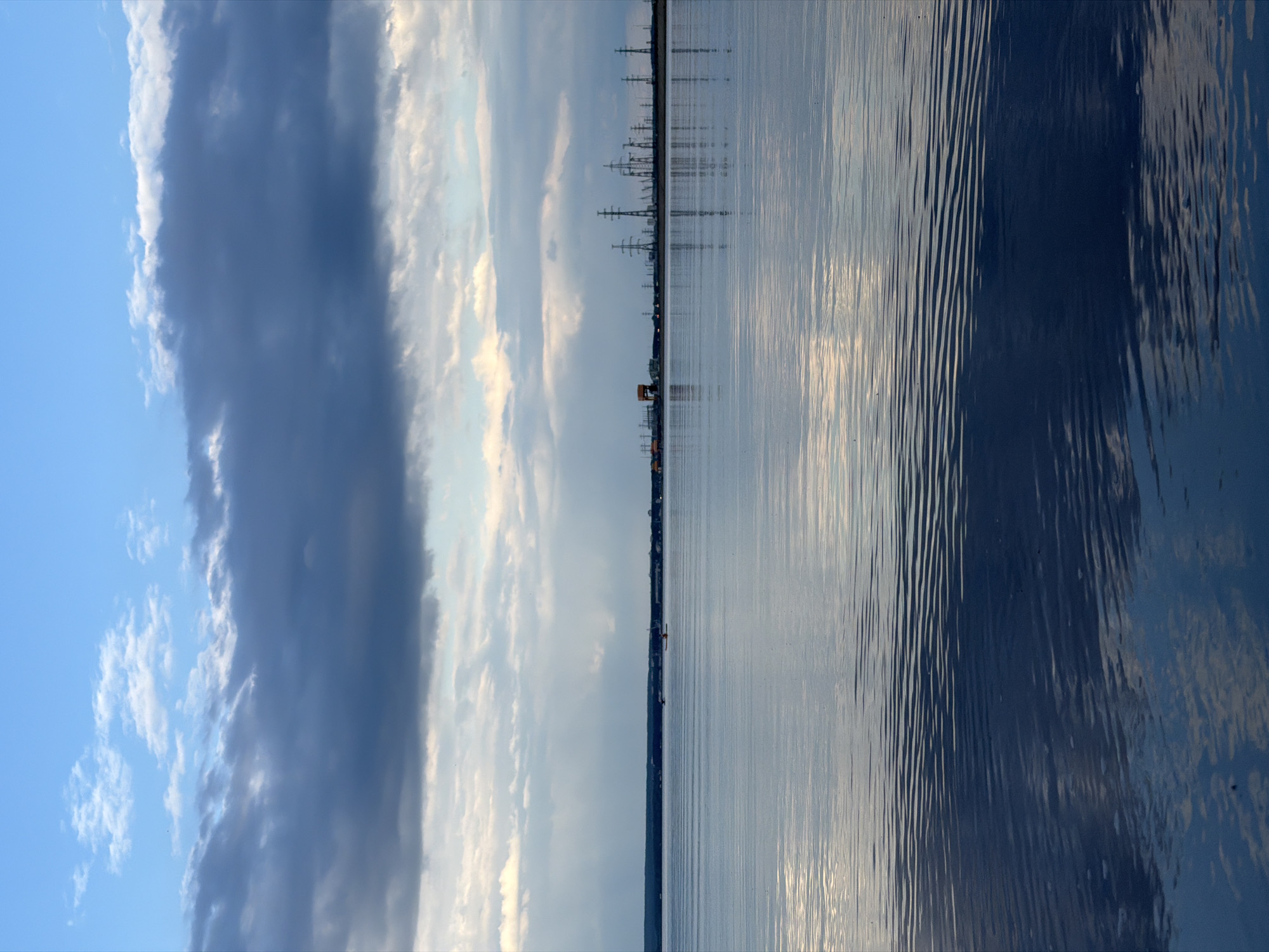 This is a photograph taken from the water's edge, looking out towards a distant industrial skyline and a cloudy sky. The water in the foreground is calm with subtle ripples reflecting the sky and clouds. Two small figures in kayaks can be seen on the water in the middle ground, far from the shore. The horizon is marked by a dark line of land, with a silhouette of numerous power pylons and some buildings visible. The sky is filled with a mix of dark, brooding clouds and lighter, wispy formations, with some shafts of sunlight breaking through. The overall impression is one of a serene, yet somewhat industrial, landscape. The location is identified as Noviy, Russia.