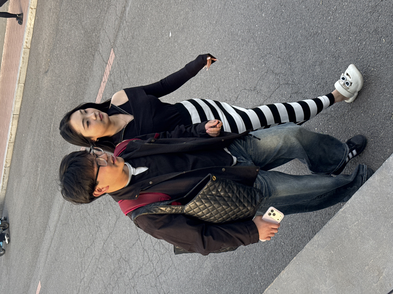 The image depicts two young adults, a man and a woman, resting on an asphalt surface in an outdoor urban setting, likely in Zhongtun, China. The perspective is from above, looking down at the subjects.
The woman is lying on her back, her long dark hair fanned out on the ground. She wears a black off-shoulder top and high-waisted black and white horizontally striped pants. Her left arm is extended towards the top of the frame. On her feet are distinctive white clogs featuring black embellishments. She gazes upwards with a serene and composed expression.
Beside her, a man is seated upright, wearing glasses, a black jacket layered over a maroon hooded top and a white undershirt. He is dressed in loose-fitting blue jeans and black sneakers with white accents. A black quilted cross-body bag is slung across his chest. He holds a light pink smartphone with a triple camera array in his right hand. His gaze is directed slightly upwards, appearing calm and relaxed.
The ground beneath them is asphalt, showing minor cracks and a faded red painted line. A concrete curb is visible along the lower and left edges of the frame, suggesting the scene is adjacent to a sidewalk or structure. The lighting indicates a clear day, possibly late afternoon, casting soft shadows. Both individuals appear to be in a casual, relaxed pose, perhaps resting or captured in a candid moment. No discernible text is visible on their clothing or the immediate surroundings.