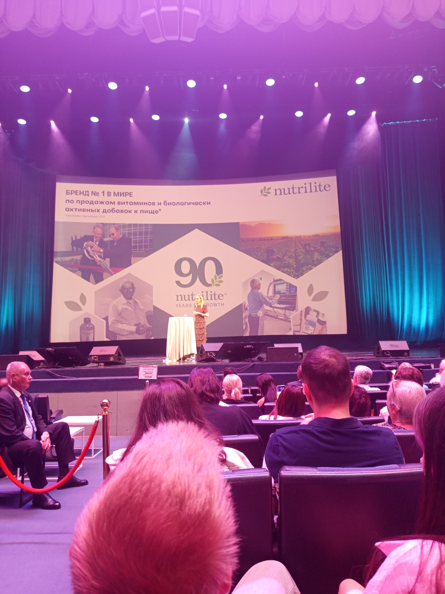 The image depicts an indoor event, possibly a conference or presentation, in Saint Petersburg, Russia. A large screen at the front displays the Nutrilite logo and text in Russian that translates to "BRAND No. 1 IN THE WORLD FOR THE SALE OF VITAMINS AND BIOLOGICALLY ACTIVE DIETARY SUPPLEMENTS." The screen also shows images related to the brand, including what appear to be people involved in the company's history or operations, and a farm landscape. A woman is speaking at a podium in front of the screen, while an audience is seated in rows of chairs facing the stage. In the foreground, a security guard in a suit is seated, and several audience members are visible from behind. The overall lighting is dim, with spotlights illuminating the stage and screen, and a pinkish-purple hue cast by ambient lighting. The venue appears to be a theater or auditorium, with curtains on the sides of the stage and decorative lighting fixtures above.