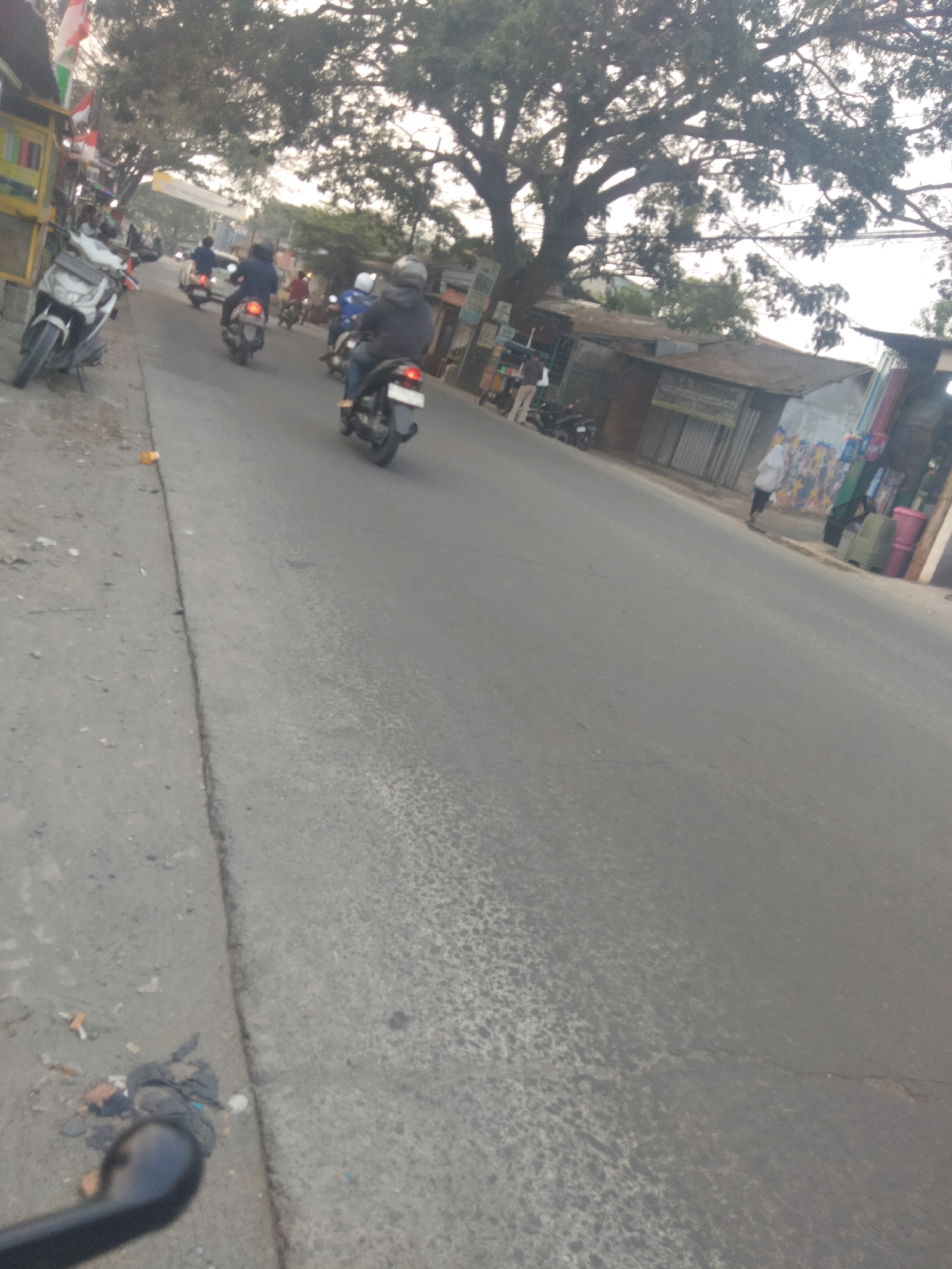 The image shows a street scene in Pisangan Jaya, Indonesia, with several motorcycles and riders. On the left side of the road, a white motorcycle is parked. Further down the road, multiple motorcycles are in motion. One rider in a dark jacket and helmet is on a motorcycle, and another rider with a helmet is behind them. A person in a blue jacket and helmet is on a motorcycle further up the street, followed by another person on a motorcycle. The environment appears to be a street with shops and trees lining the sides. The road surface is asphalt with some wear and tear. The lighting suggests it is daytime, possibly late afternoon or early morning, with some hazy conditions. Trash is visible on the side of the road in the foreground.