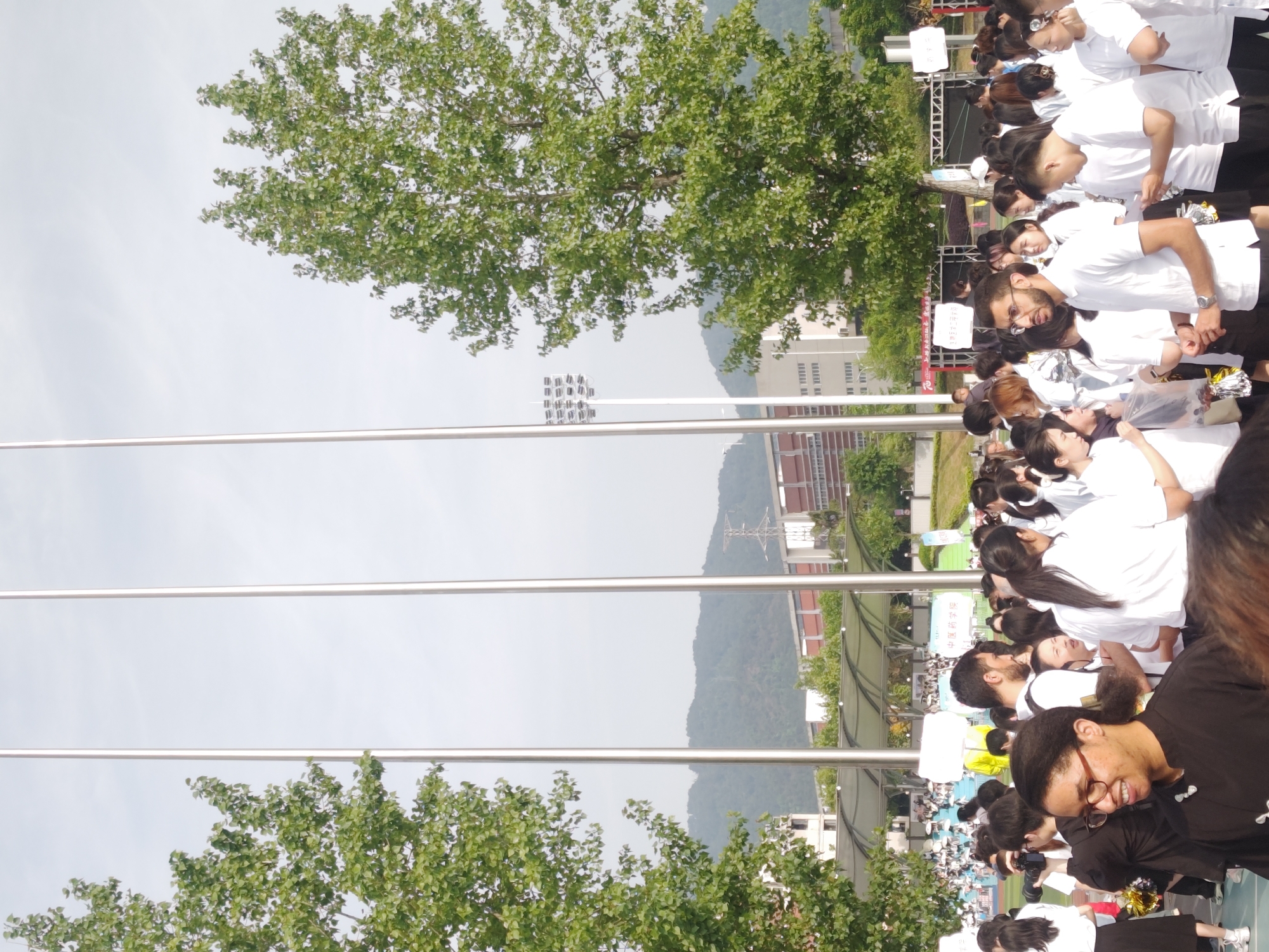 A medium shot shows a crowded outdoor scene in Chongqing High-tech Zone, China. People, primarily dressed in white shirts, are interacting and facing the right. There are metal poles, trees with green leaves, mountains in the background, and buildings. A light tower is visible.