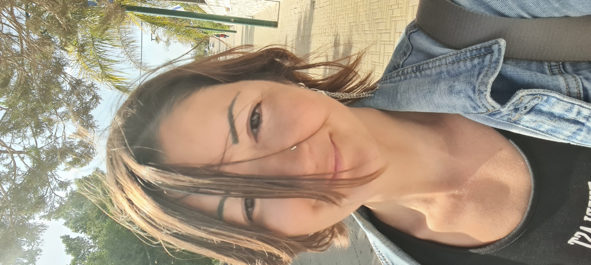 The image is an outdoor selfie, horizontally oriented, featuring a woman in a bright, sunny environment, likely in the Lachish Regional Council, Israel. The woman, appearing to be in her 20s or 30s, is positioned in the foreground, looking directly at the camera with a relaxed, slight smile. She has medium-length brown hair that is windswept, partially obscuring her face. Her dark eyes and defined eyebrows are visible, along with a small silver nose piercing and a silver chain necklace.
She is wearing a light-wash denim jacket over a black top. Partial white text is visible on the black top, but it is fragmented and unreadable. The bright daylight highlights her features and suggests a clear day.
The background shows an outdoor setting with ample greenery. Various trees, including palm trees and other foliage, are visible against a light blue sky. Below the trees, there is a paved area, likely a sidewalk or street, featuring linear patterns. In the distance, a light-colored building or wall is present, with shadows indicating strong sunlight. The overall scene conveys a pleasant, relaxed atmosphere.